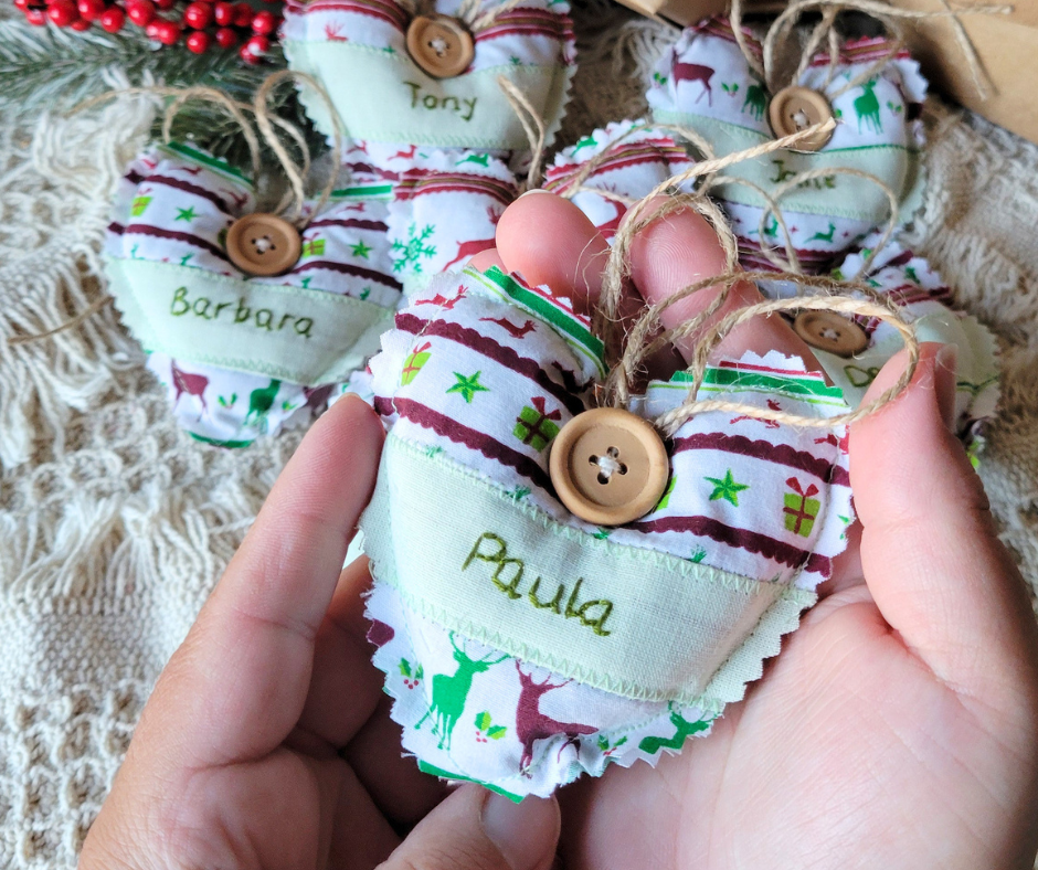 Hands holding a fabric heart filled with lavender and embroidered on the front is the name Paula. The heart is white with green and burgundy colours and has reindeer on it. In the background are similar hearts with different names.