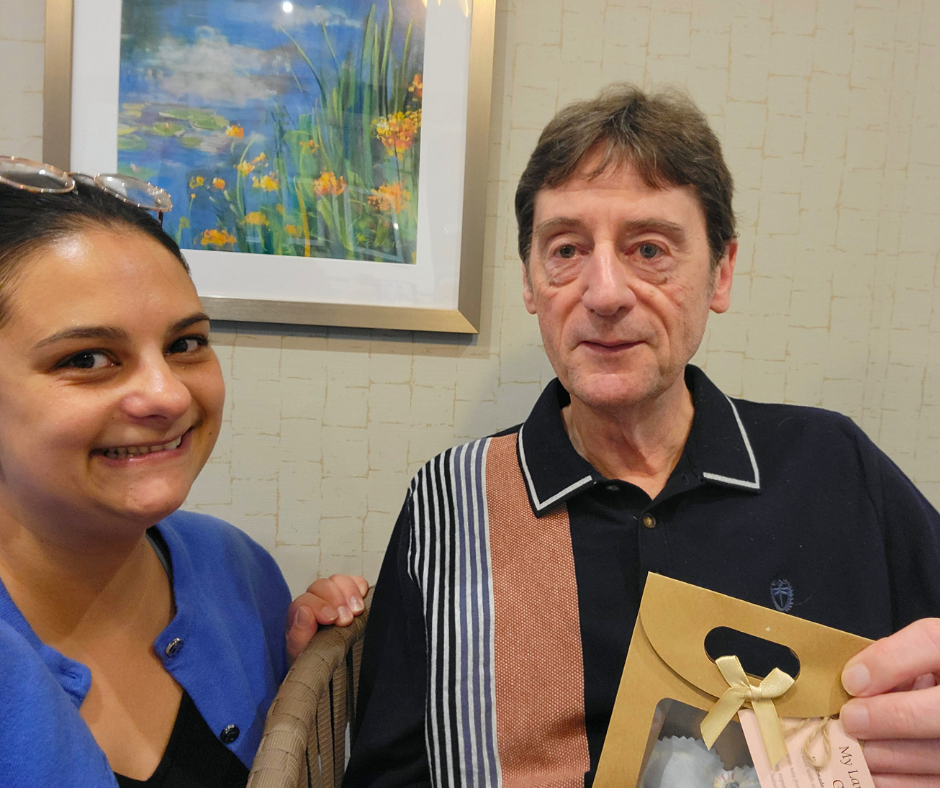 A woman smiling, wearing sunglasses on her head and a blue shirt, standing next to an older man holding a gift bag. They are indoors with a framed floral painting on the beige wall behind them.