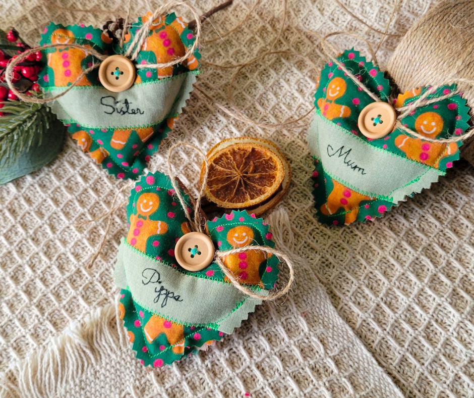 Festive fabric heart-shaped Christmas decorations labeled 'Sister,' 'Mum,' and 'Pippa,' with buttons and twine, arranged with a dried orange slice on a textured beige fabric surface.