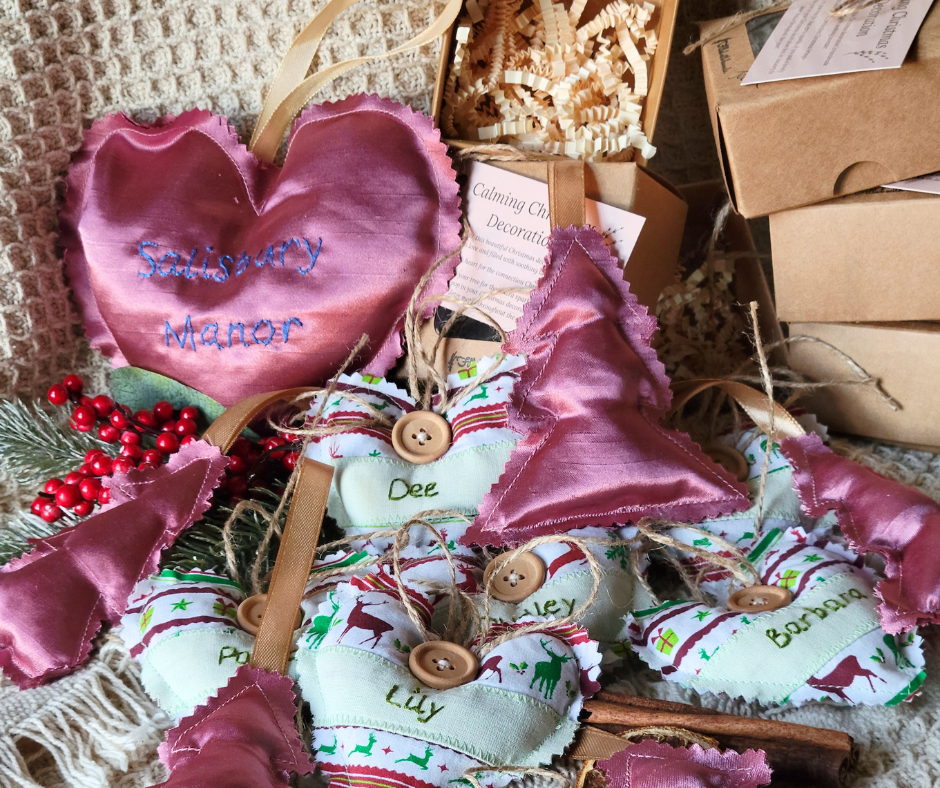 Assorted handmade fabric Christmas decorations, including pink satin heart embroidered with names, fabric hearts with Christmas themed fabrics, a sprig of red berries with pine needles, and boxes in the background.