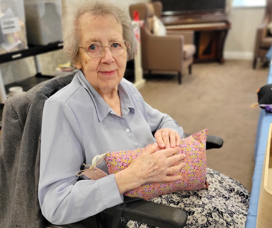 Elderly women with grey hair, glasses and blue shirt is sat in a chair holding a pink cushion