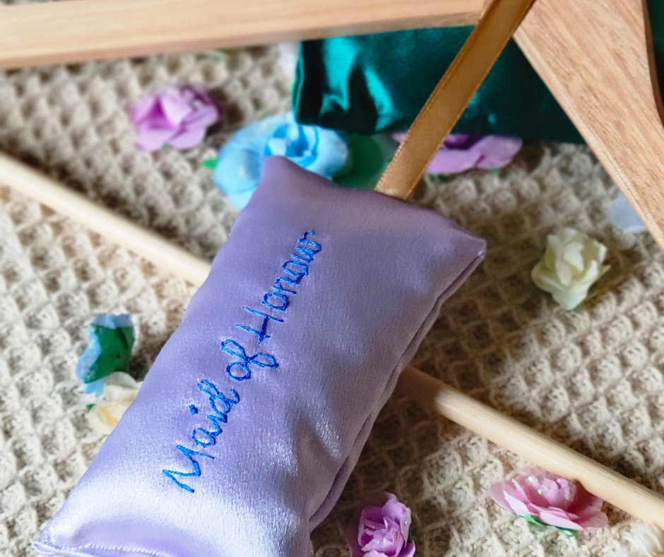 A purple fabric pouch embroidered with 'Marie of Homes' resting on a textured beige surface, surrounded by colorful flower petals and a wooden frame, beneath a piece of furniture with wooden legs.