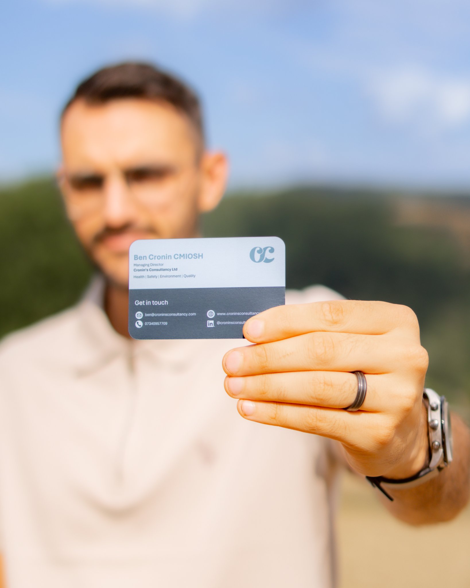 Person holding a business card outdoors, with the person's face blurred in the background. The business card displays contact information, including name, email, phone number, and website.
