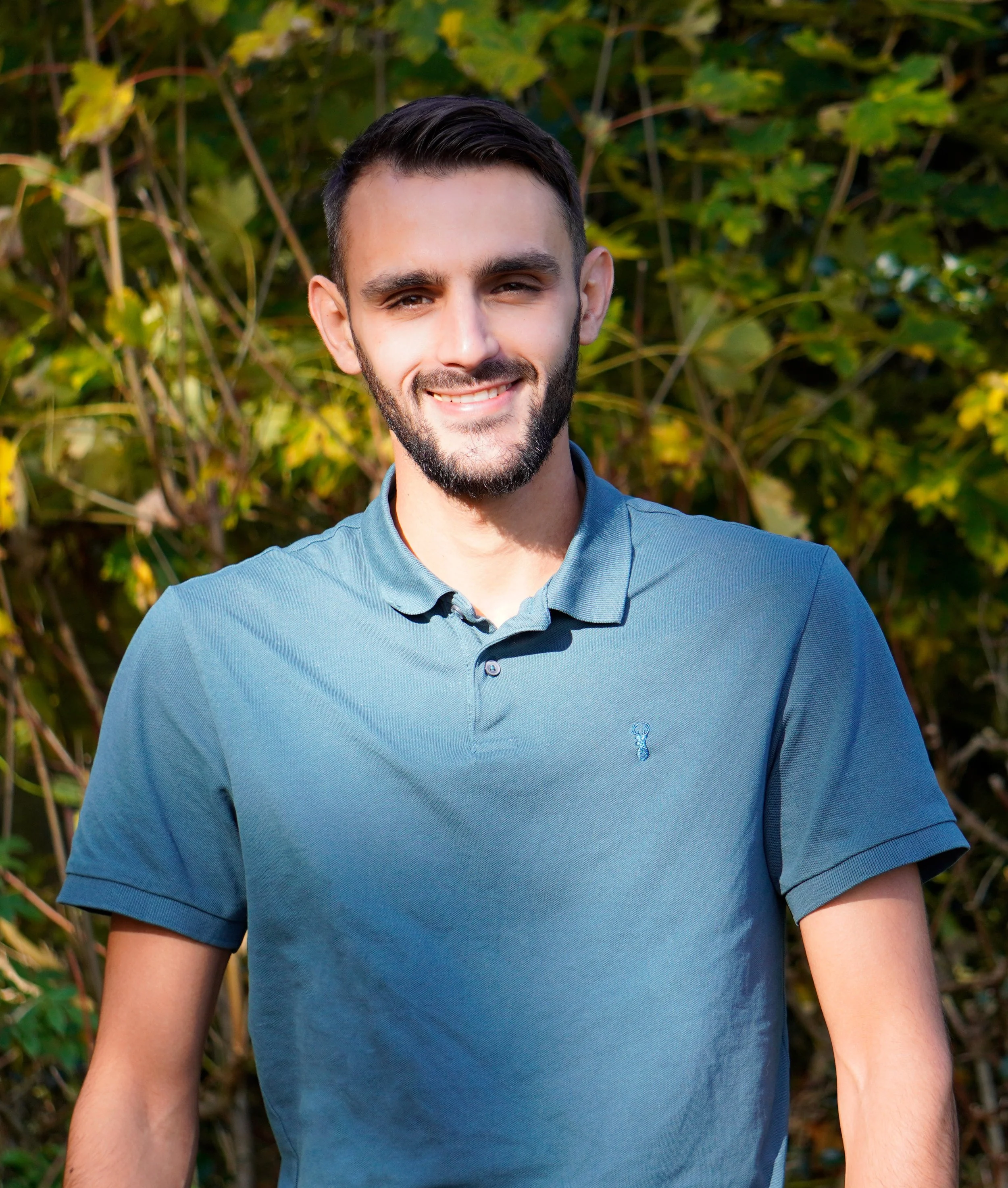 Close-up of a young man with short, dark hair and a beard, smiling in front of green leafy bushes, wearing a blue collared shirt.