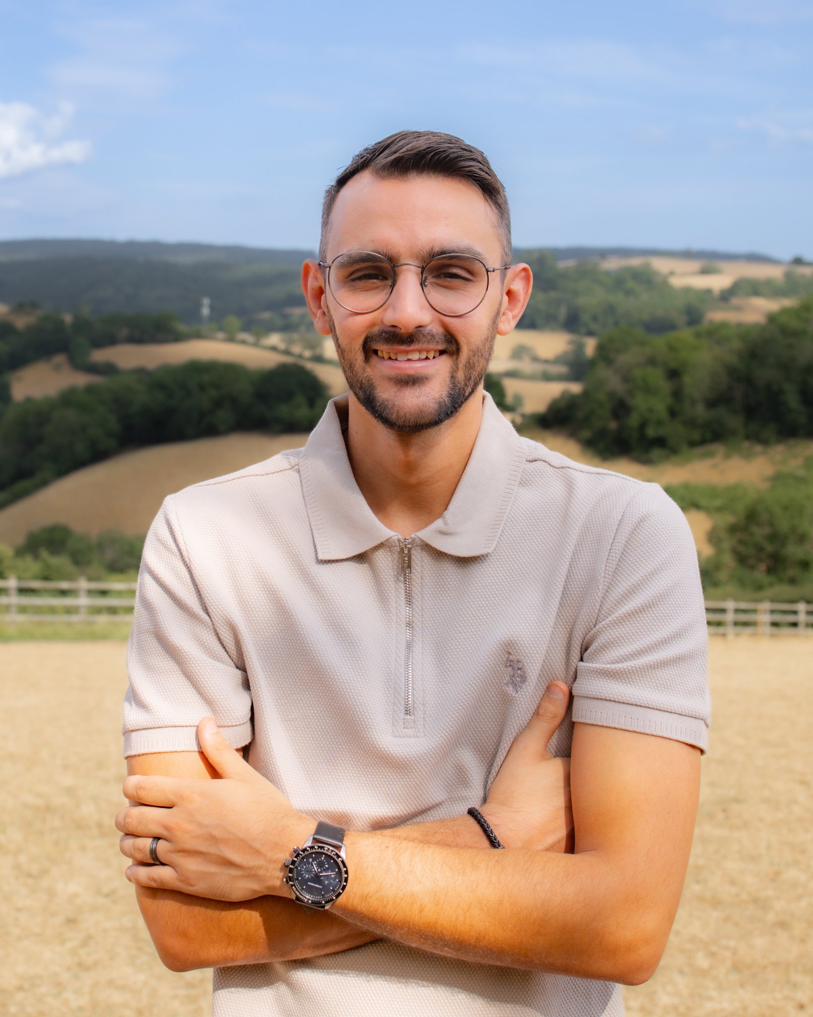 A young man with glasses and a beard stands outdoors on a sunny day with rolling hills and trees in the background. He is smiling, wearing a beige zip-up polo shirt, a wristwatch, and a bracelet. His arms are crossed.