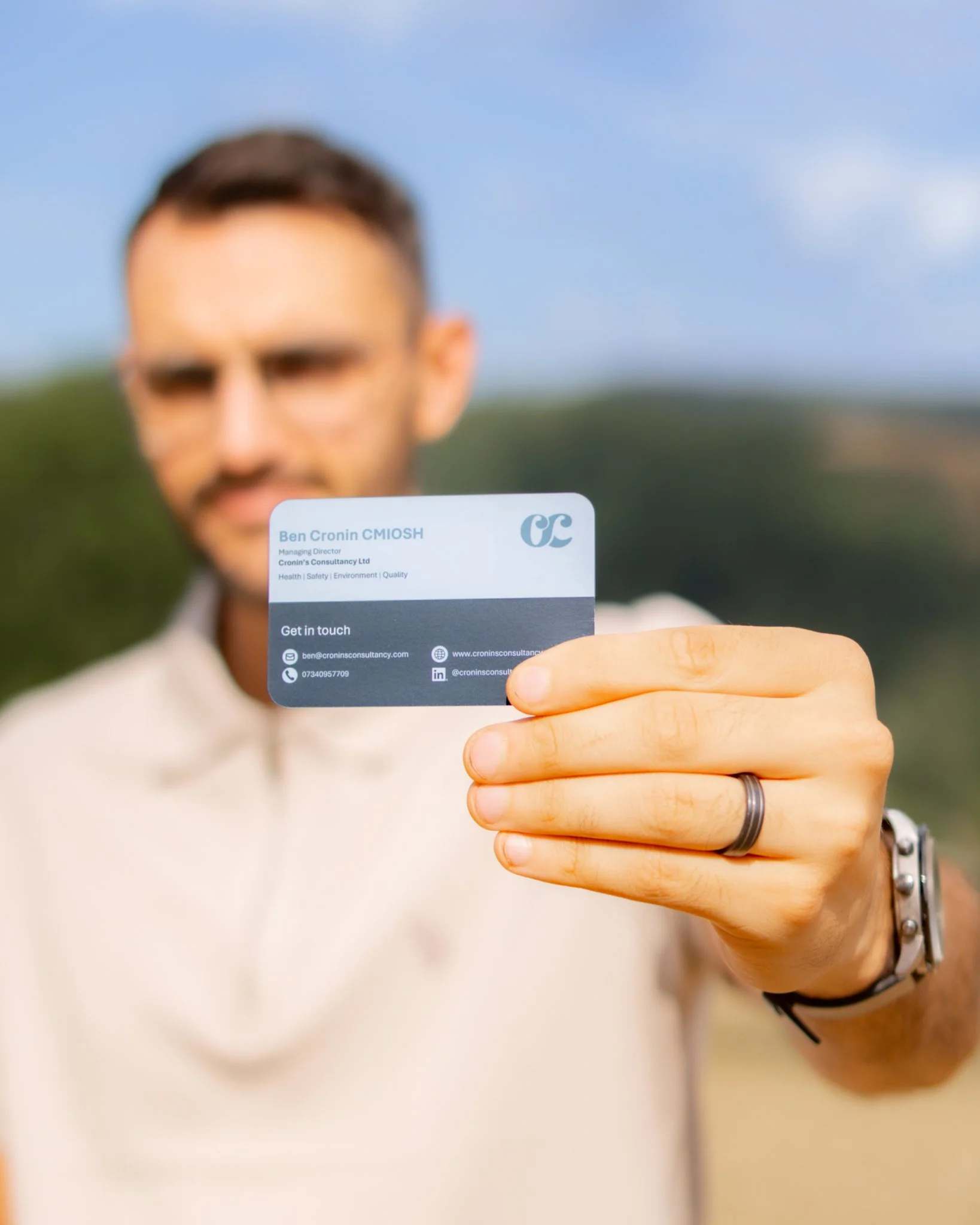 Close-up of a man's hand holding a business card with his contact information in focus, while his face is blurred in the background outdoors.