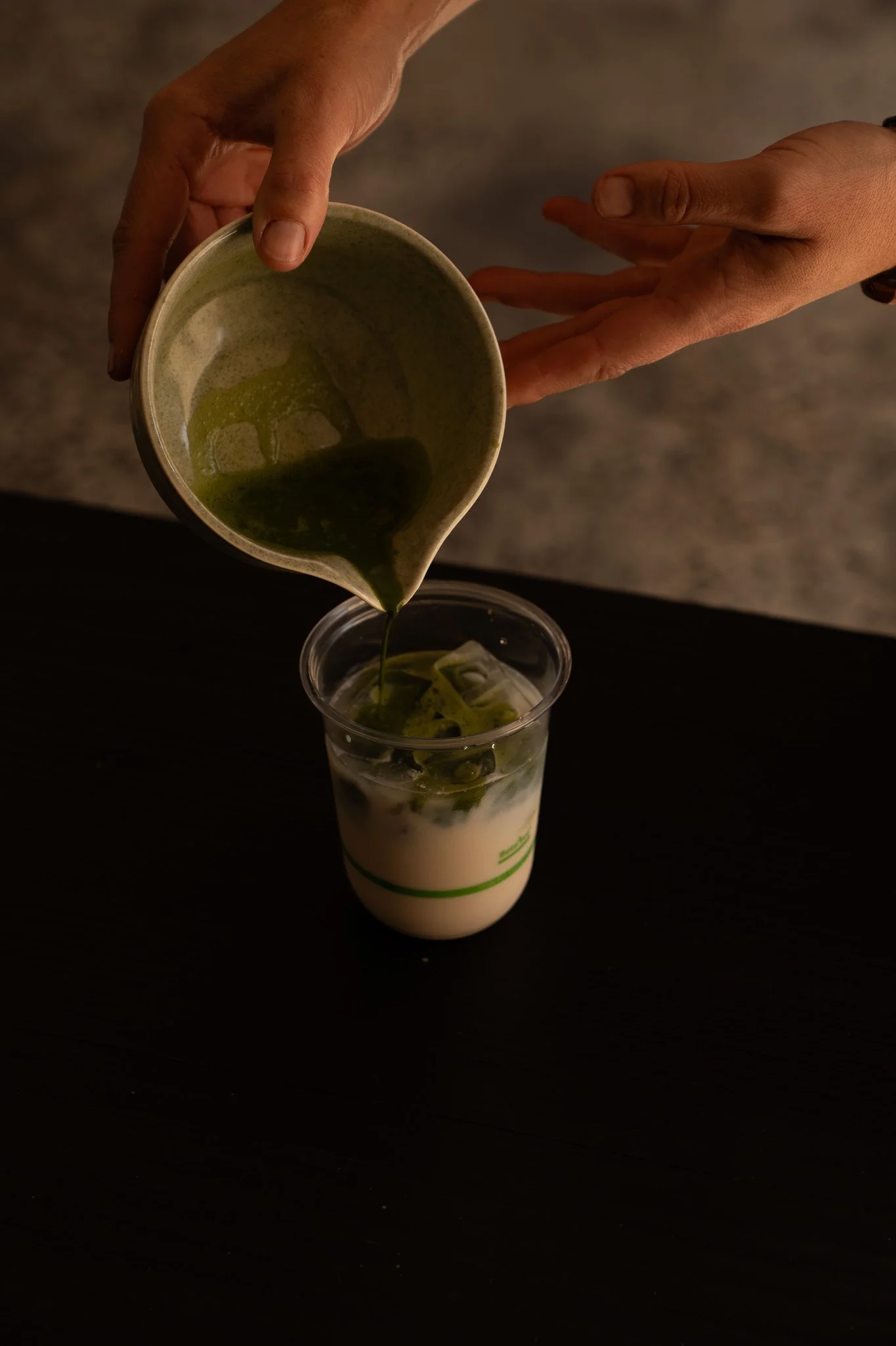 A person pours a green matcha tea mixture into a clear plastic cup filled with ice and milk in retail space at koko studio in Erina, Central Coast.