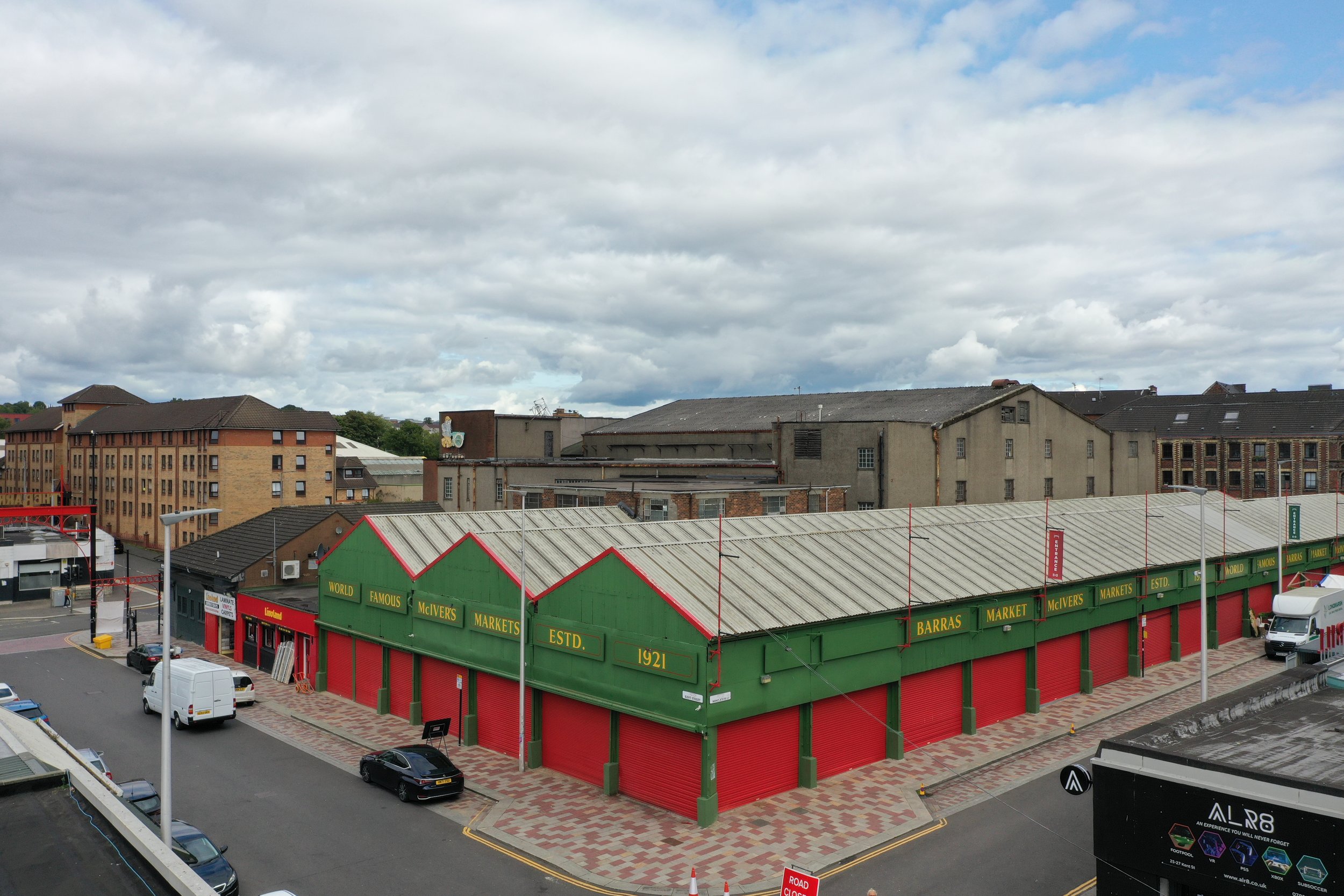 A cityscape view featuring a green and red building with a sign that reads 'McIvers Markets' in front of a brown brick building and taller structures, with a cloudy sky overhead. A bird's eye view of the Barras Market.