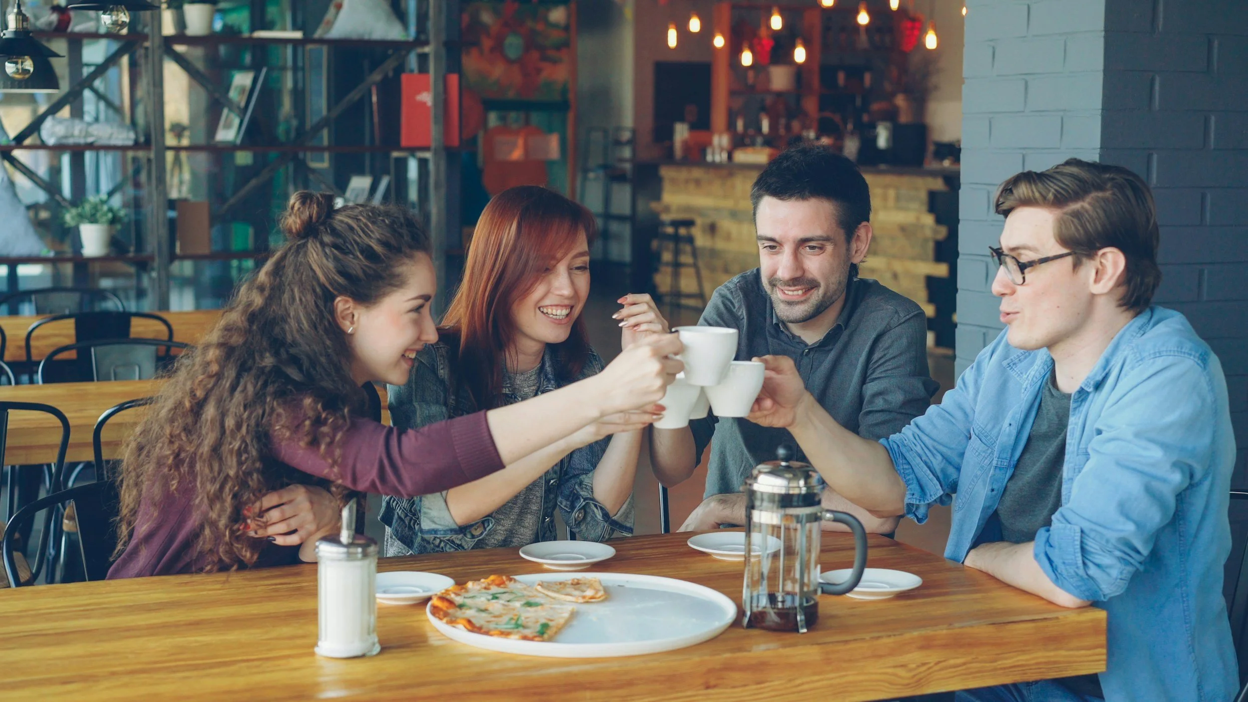 Four young adults sitting at a wooden table in a cozy cafe, smiling and toasting with coffee cups. There's a pizza on a plate and a French press on the table. The background features industrial-style decor with shelves, books, plants, and hanging lights.