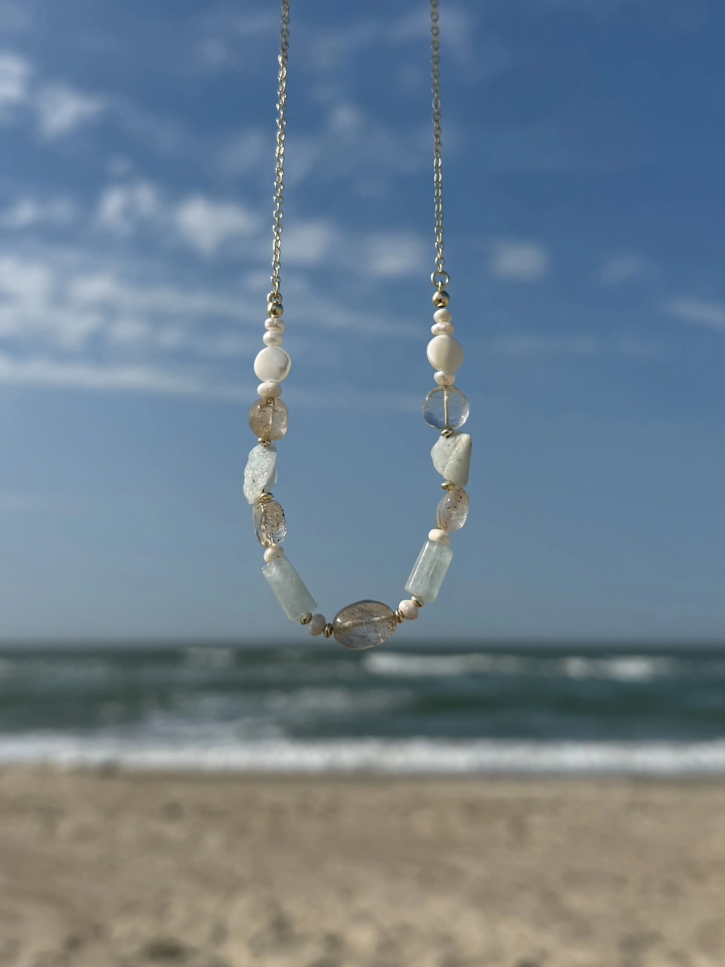 Handmade gemstone necklace with pale blue and translucent natural stones suspended against an ocean background.