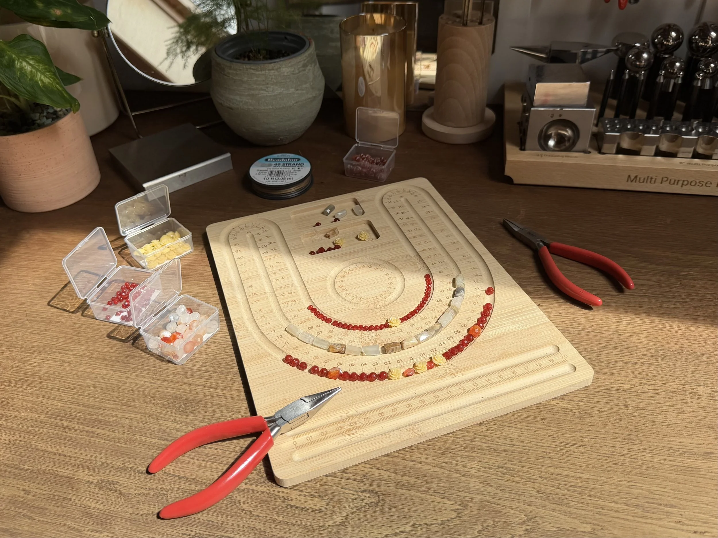 A jewelry-making workspace with a beaded necklace and various tools, containers of beads, and plants on a wooden surface.