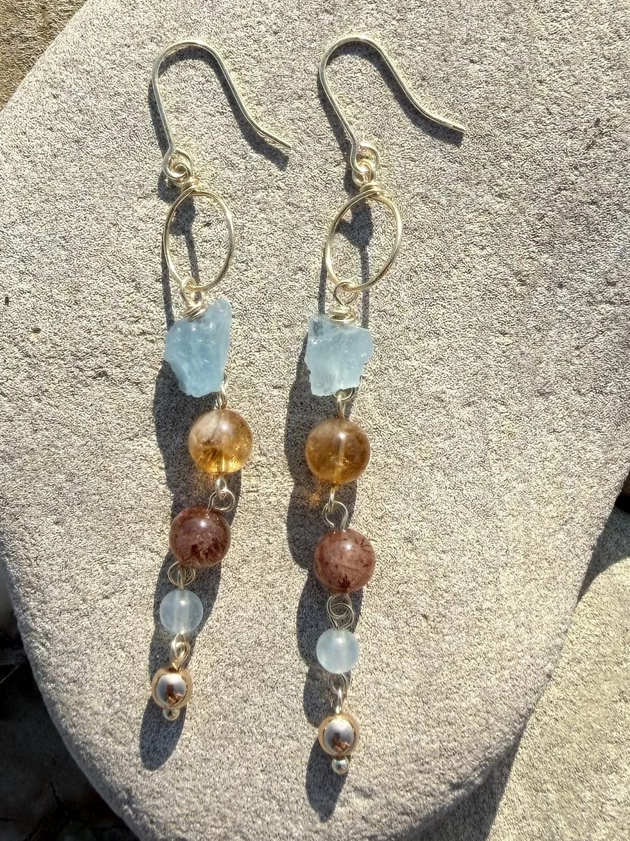 Pair of handmade gemstone drop earrings with blue, amber, brown and translucent gemstones displayed on a stone.