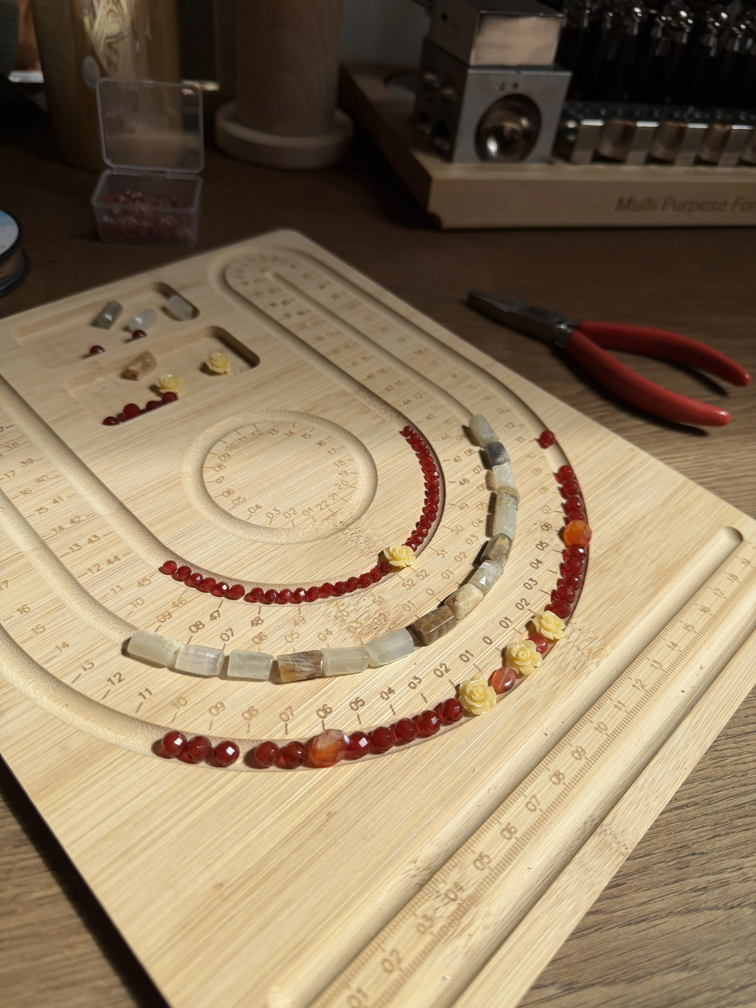 A wooden beading board with various necklaces made of semi-precious stones and beads, a pair of red-handled wire cutters, and a small plastic container with red beads on a wooden surface.