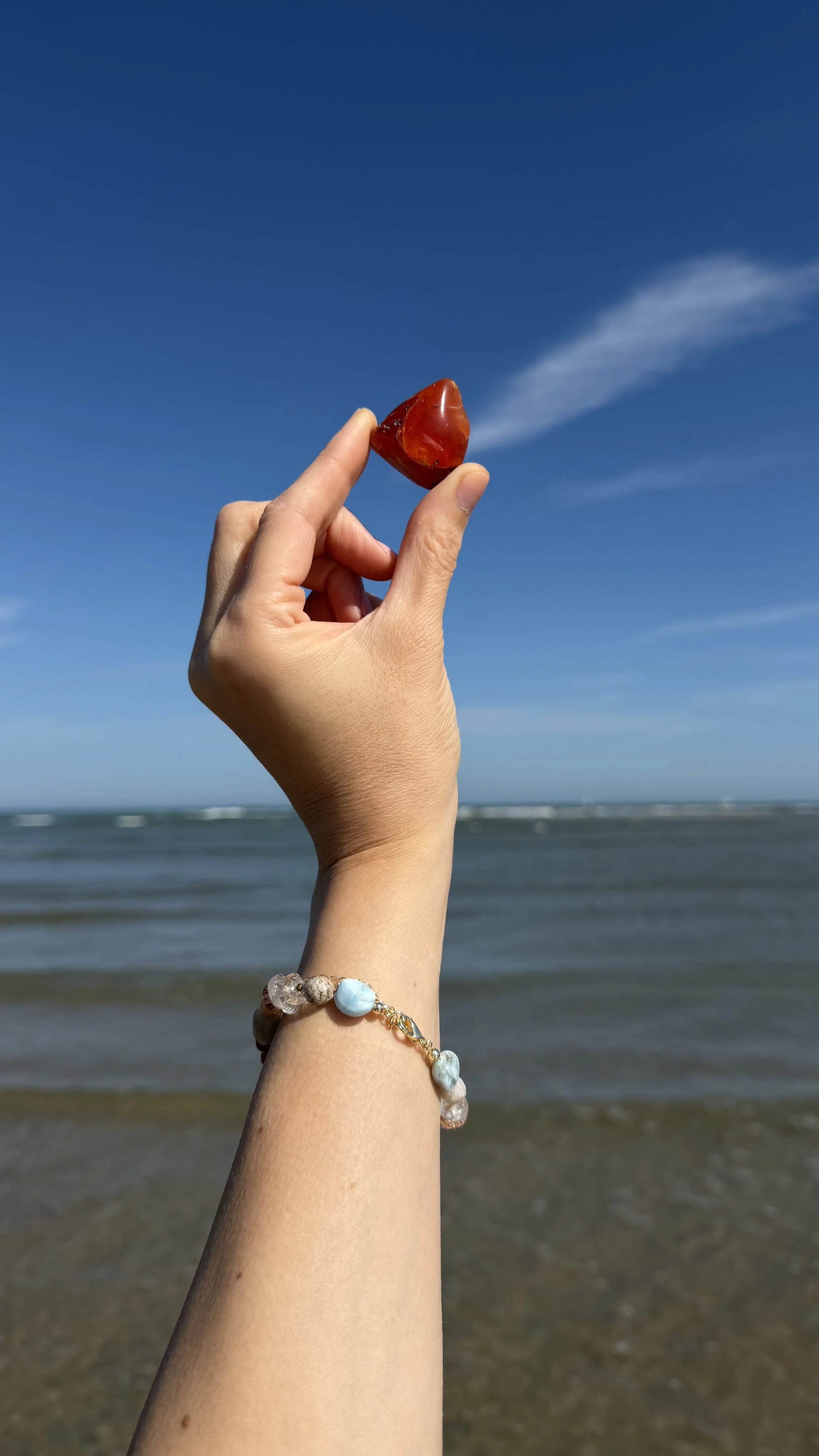 Handmade gemstone bracelet worn at the beach while holding a red stone, with the sea in the background.