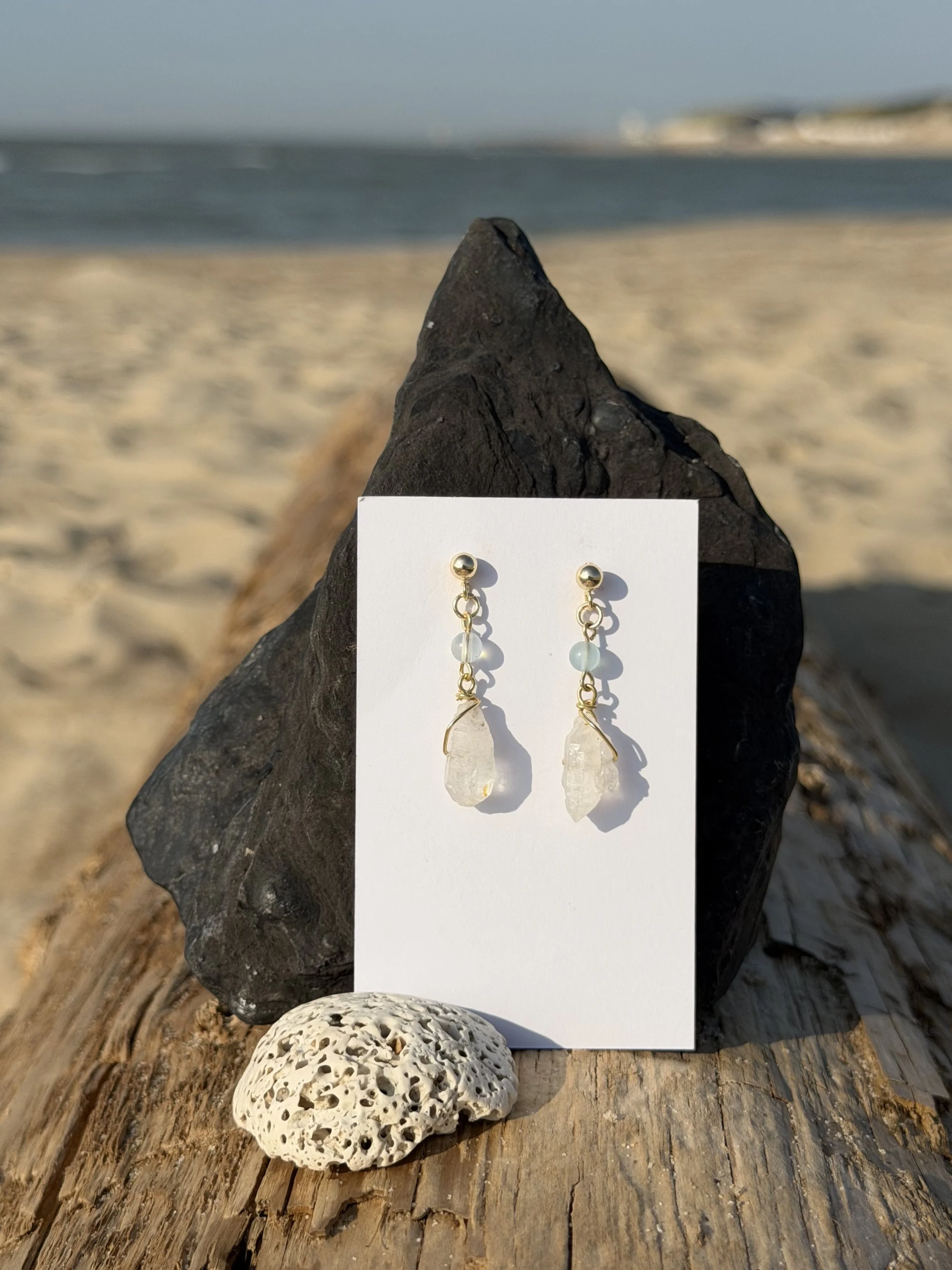 Pair of handmade gemstone drop earrings with translucent stones displayed on a card against driftwood at the beach.