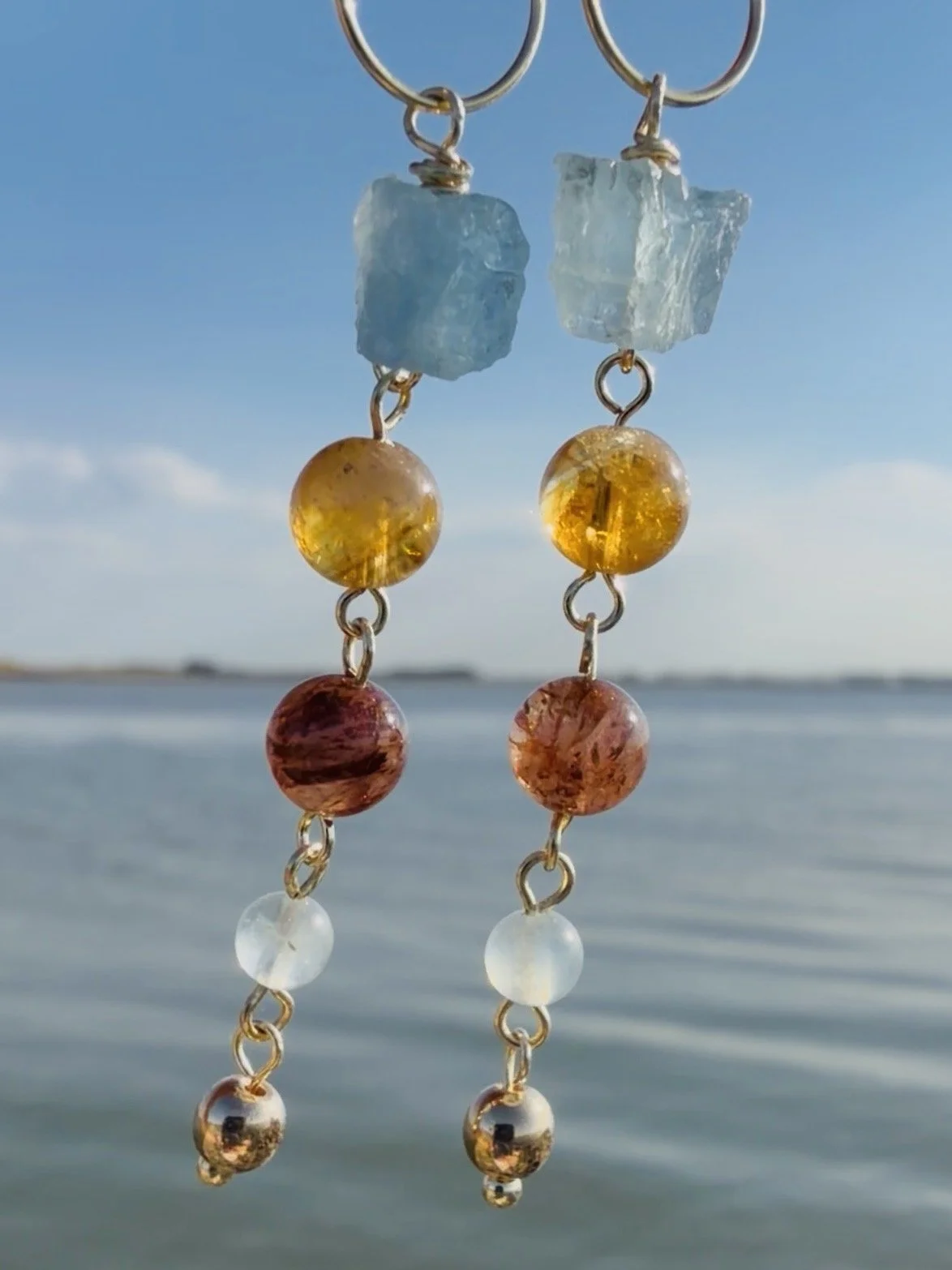 Close-up of handmade gemstone drop earrings with blue, amber, brown and translucent natural gemstones by the sea.