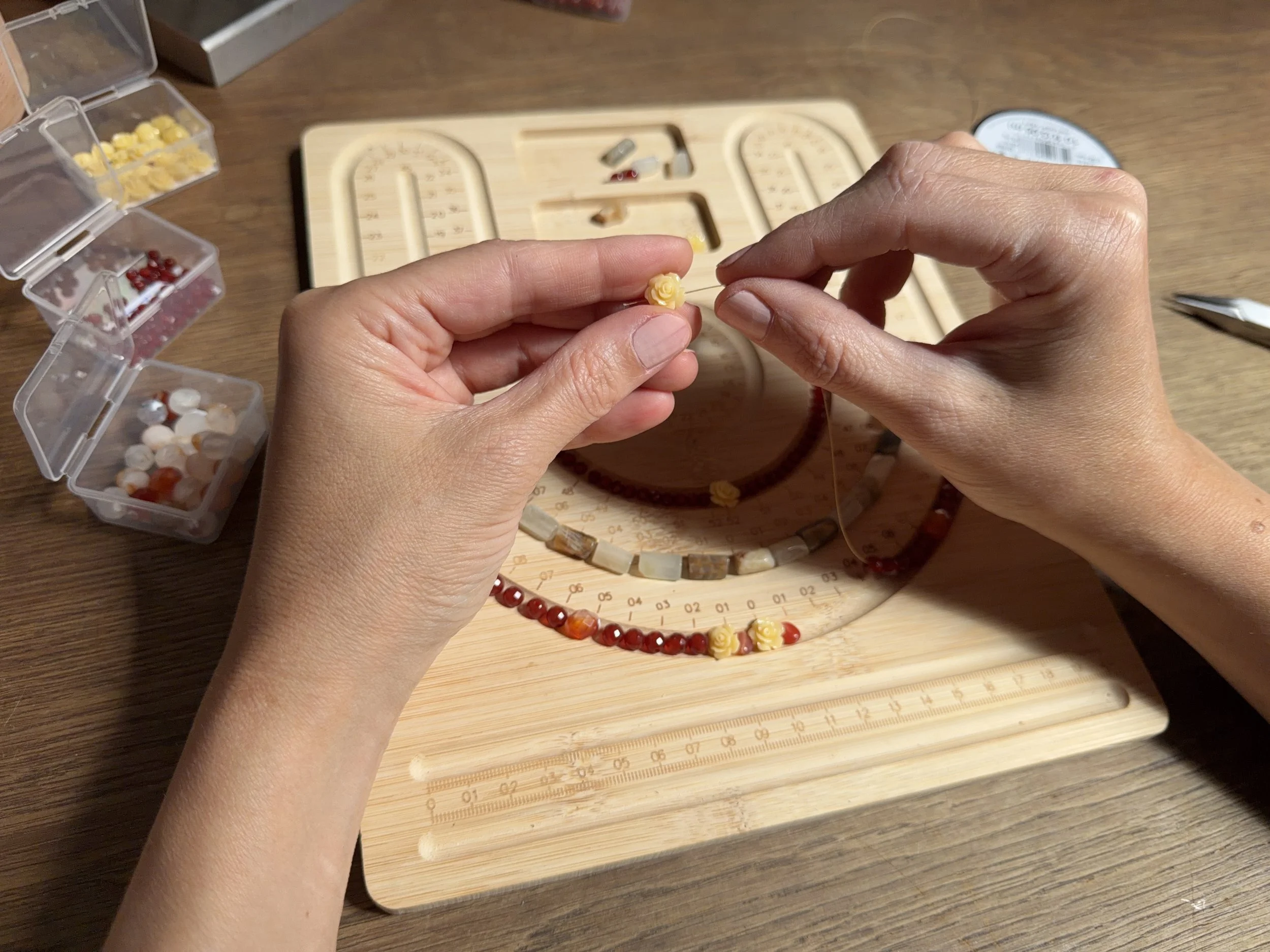 Hands sewing a beaded necklace over a wooden sewing board with measuring markings, small containers of beads nearby.