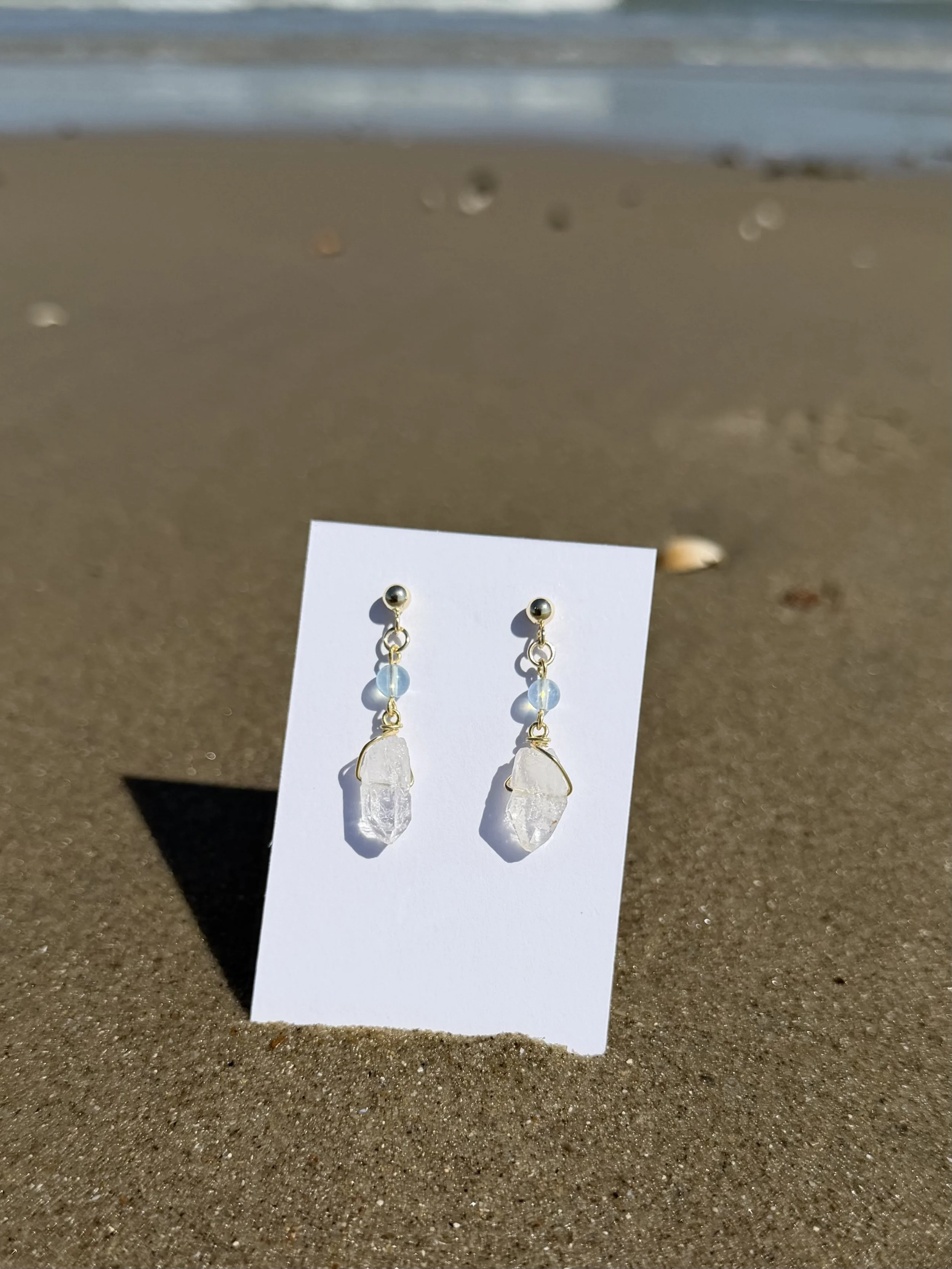 Pair of handmade gemstone drop earrings with translucent stones displayed on a card in the sand.