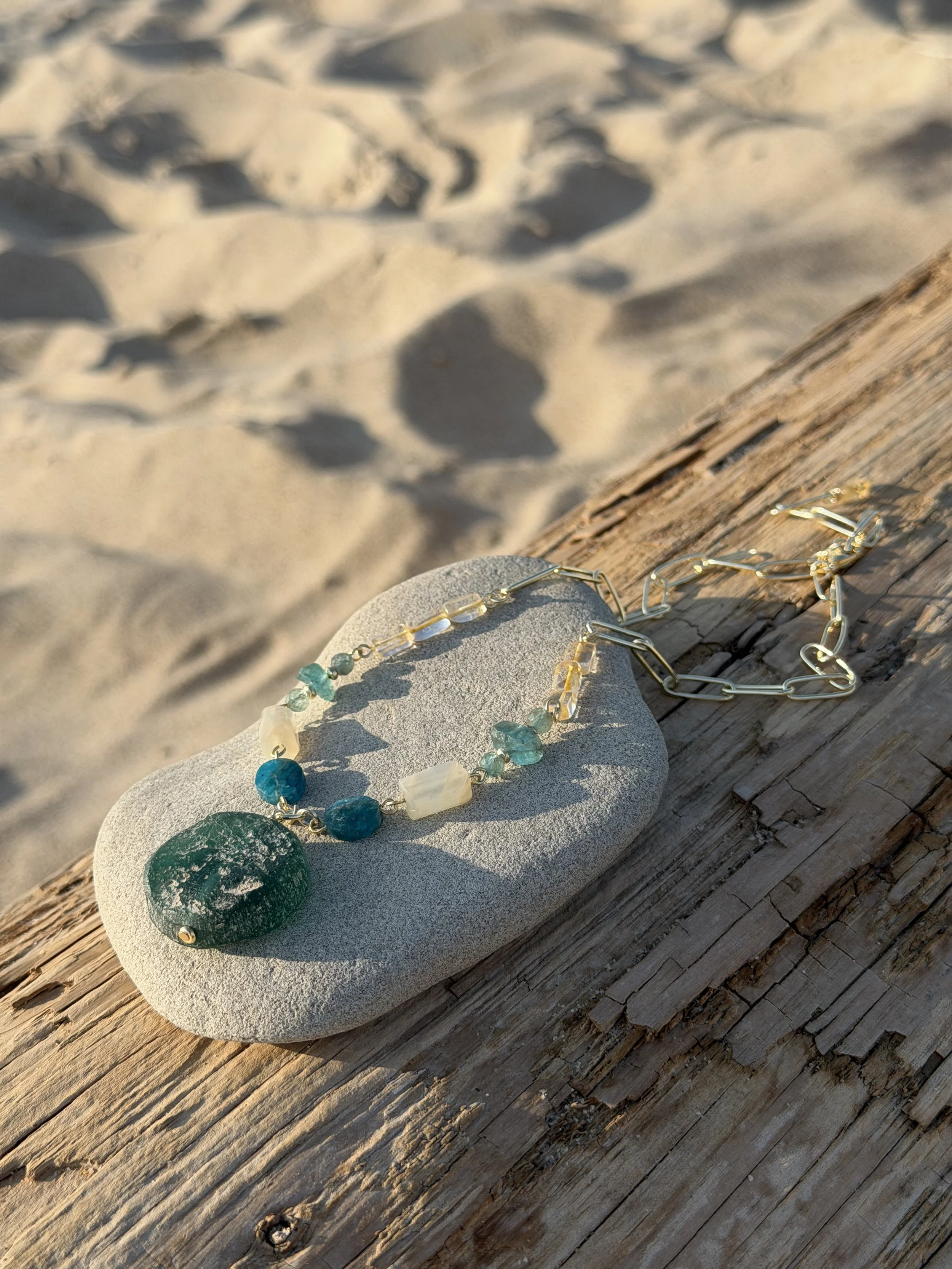 A necklace with blue, green, and clear stones resting on a gray stone and a piece of weathered wood on a sandy beach.