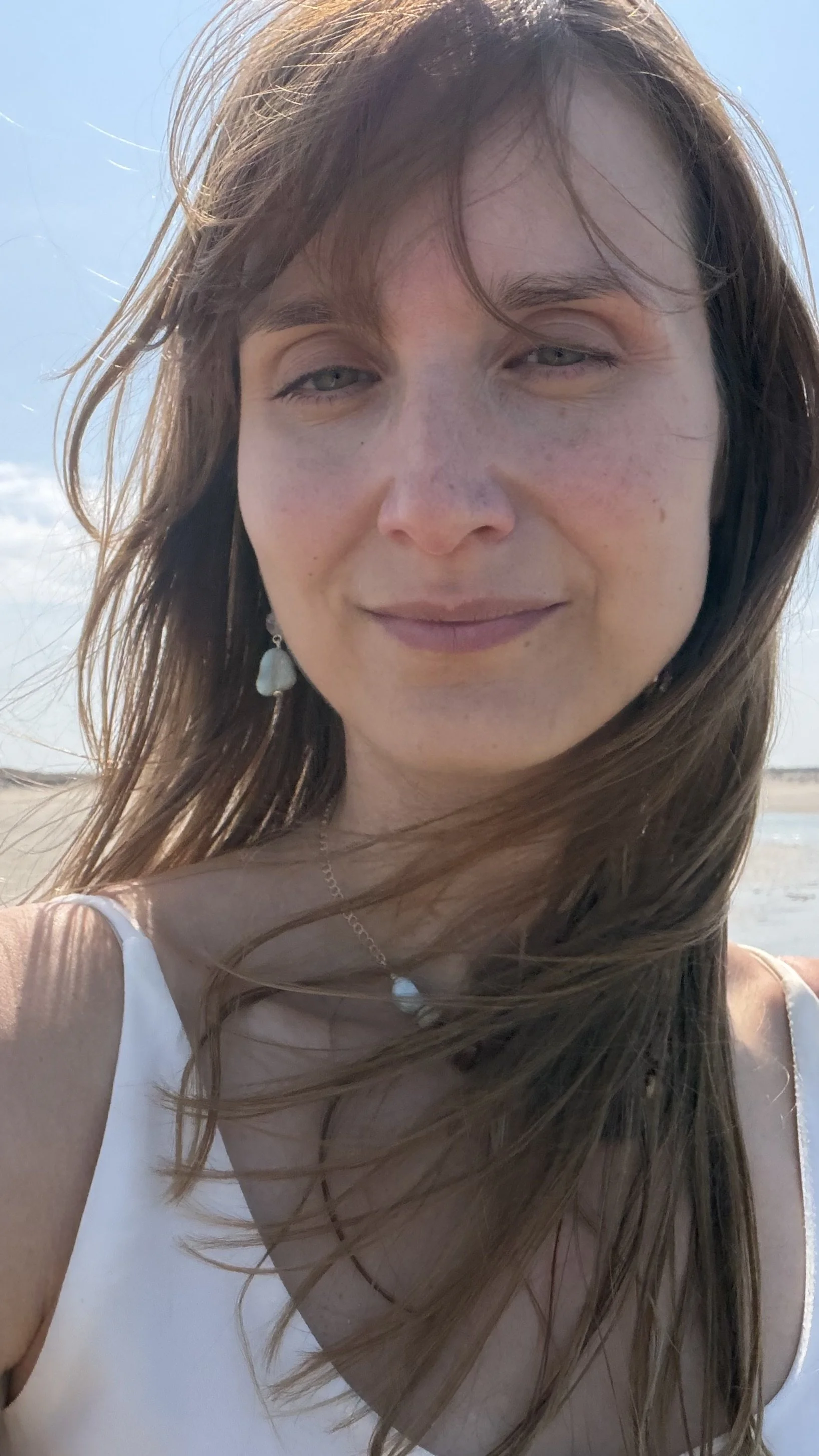 Close-up of a woman with long brown hair waving in the wind at the beach, wearing a white dress and jewelry, sunny day with blue sky.