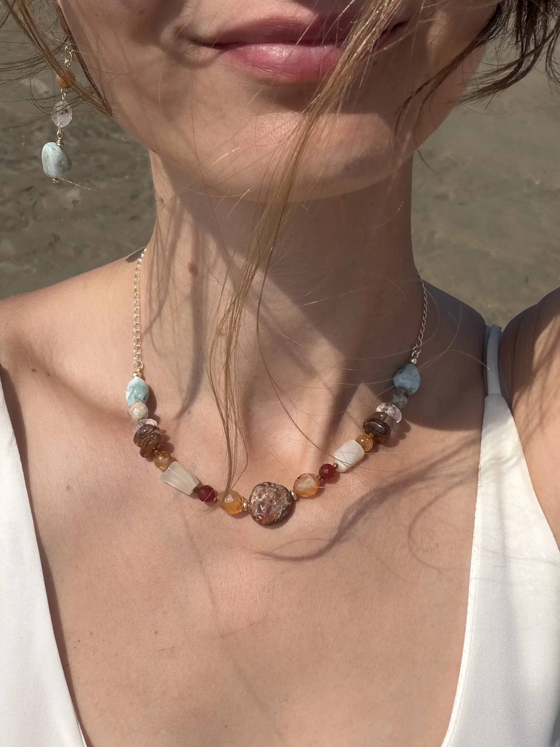 Handmade gemstone necklace with blue, cream, amber and red natural stones displayed on a smooth stone by the sea.