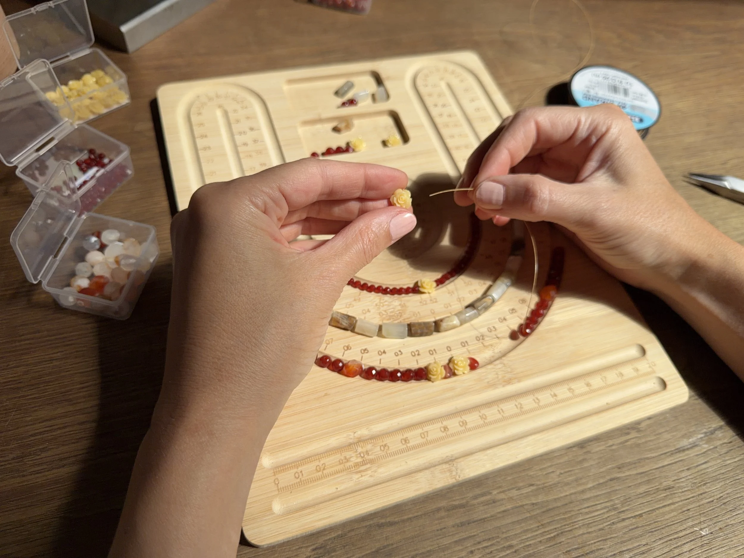 A person is working on making a beaded necklace with yellow, white, and red beads, using a bead mat and tools on a wooden table surrounded by small containers of beads.