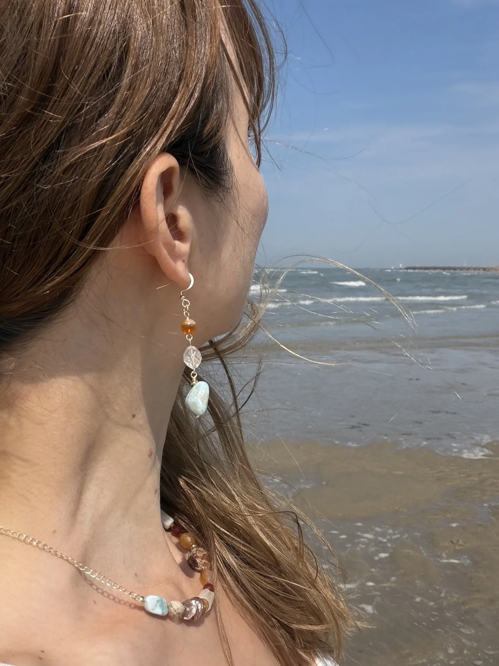 Handmade gemstone drop earring with orange, translucent and pale blue stones worn by the sea, showing its length and delicate design.