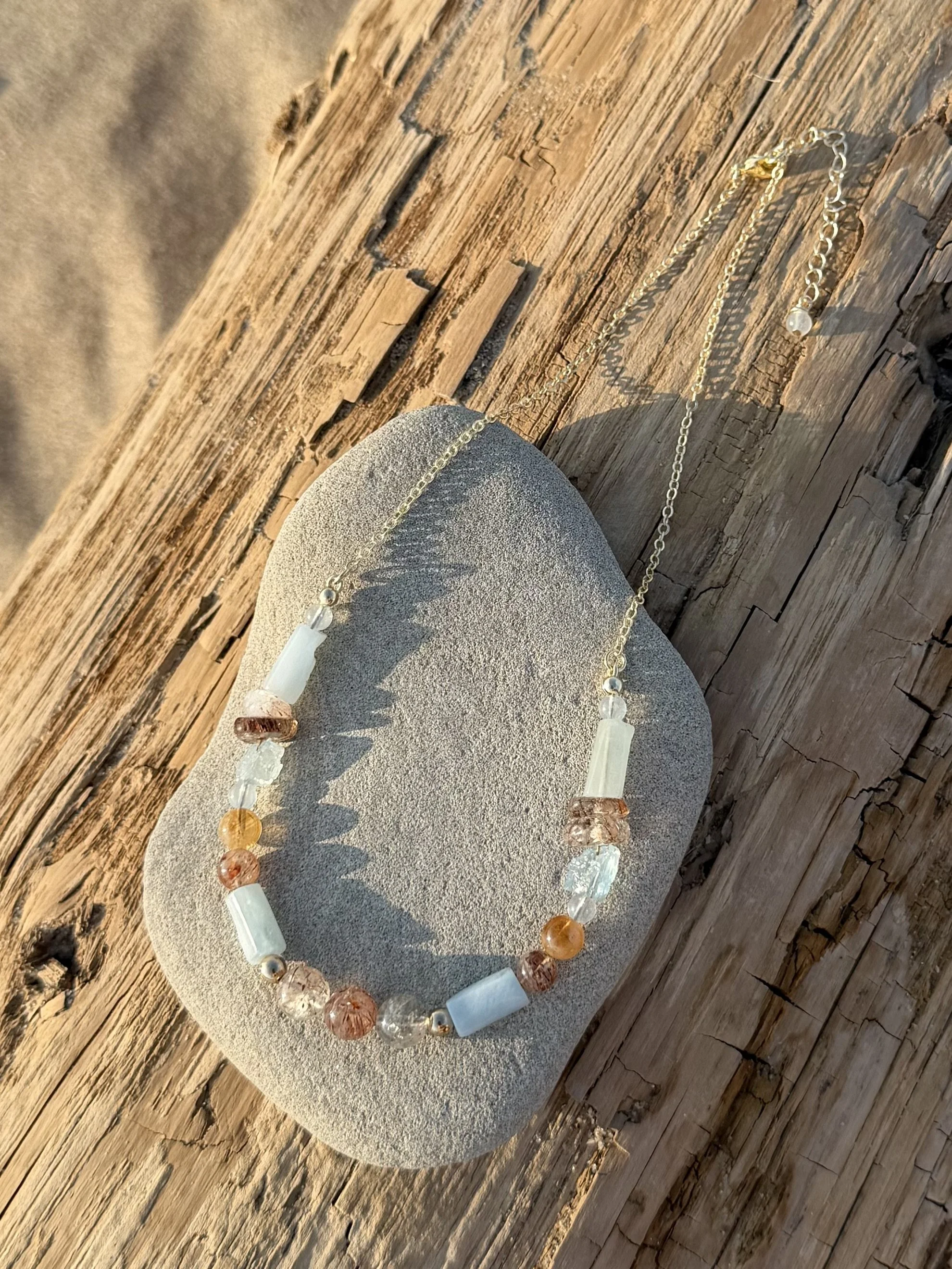 Handmade gemstone necklace with blue, white and warm-toned natural gemstones displayed on a stone and driftwood.