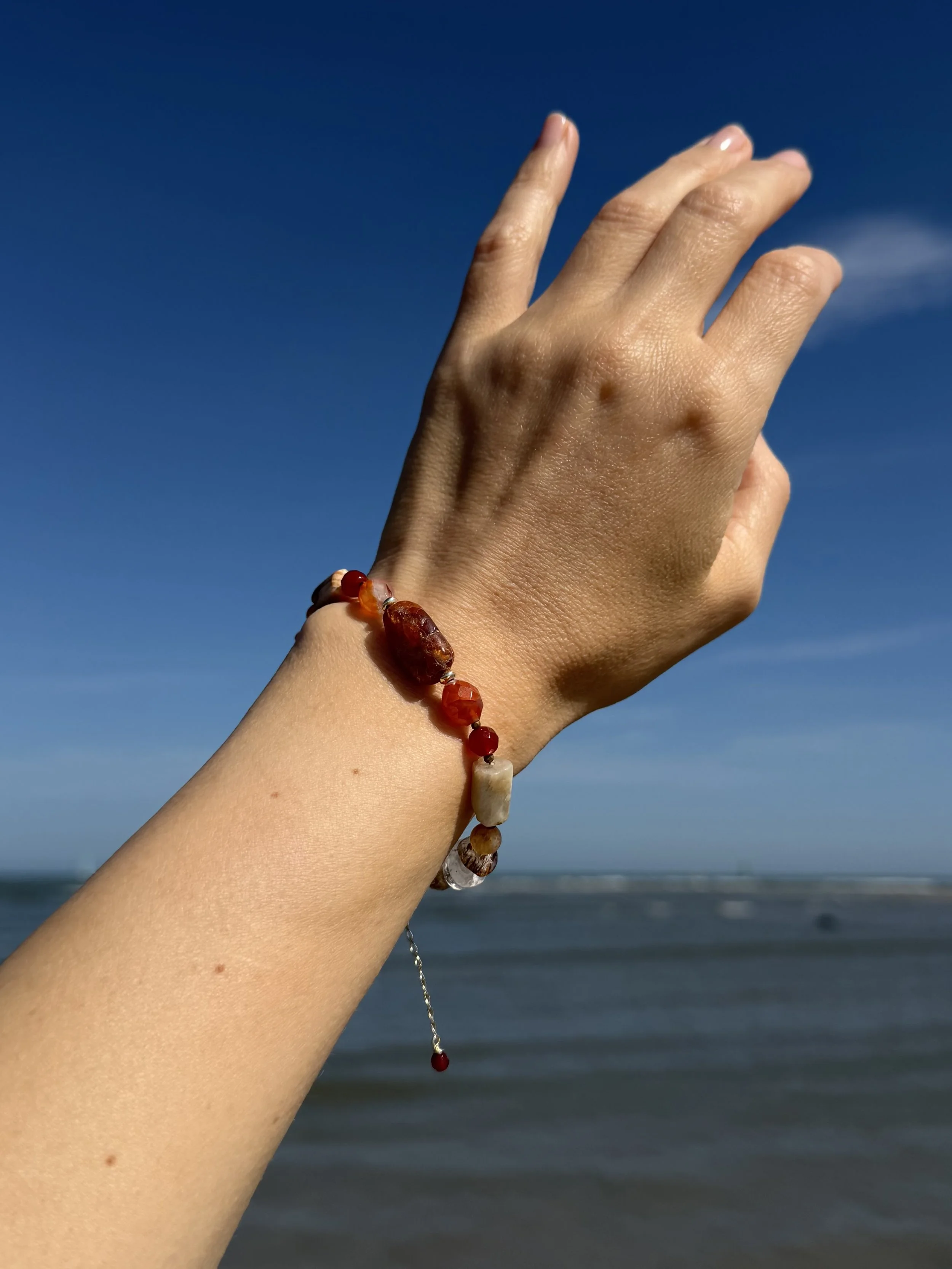 Colourful handmade gemstone bracelet worn on the wrist at the beach, showing its warm red and amber gemstones.