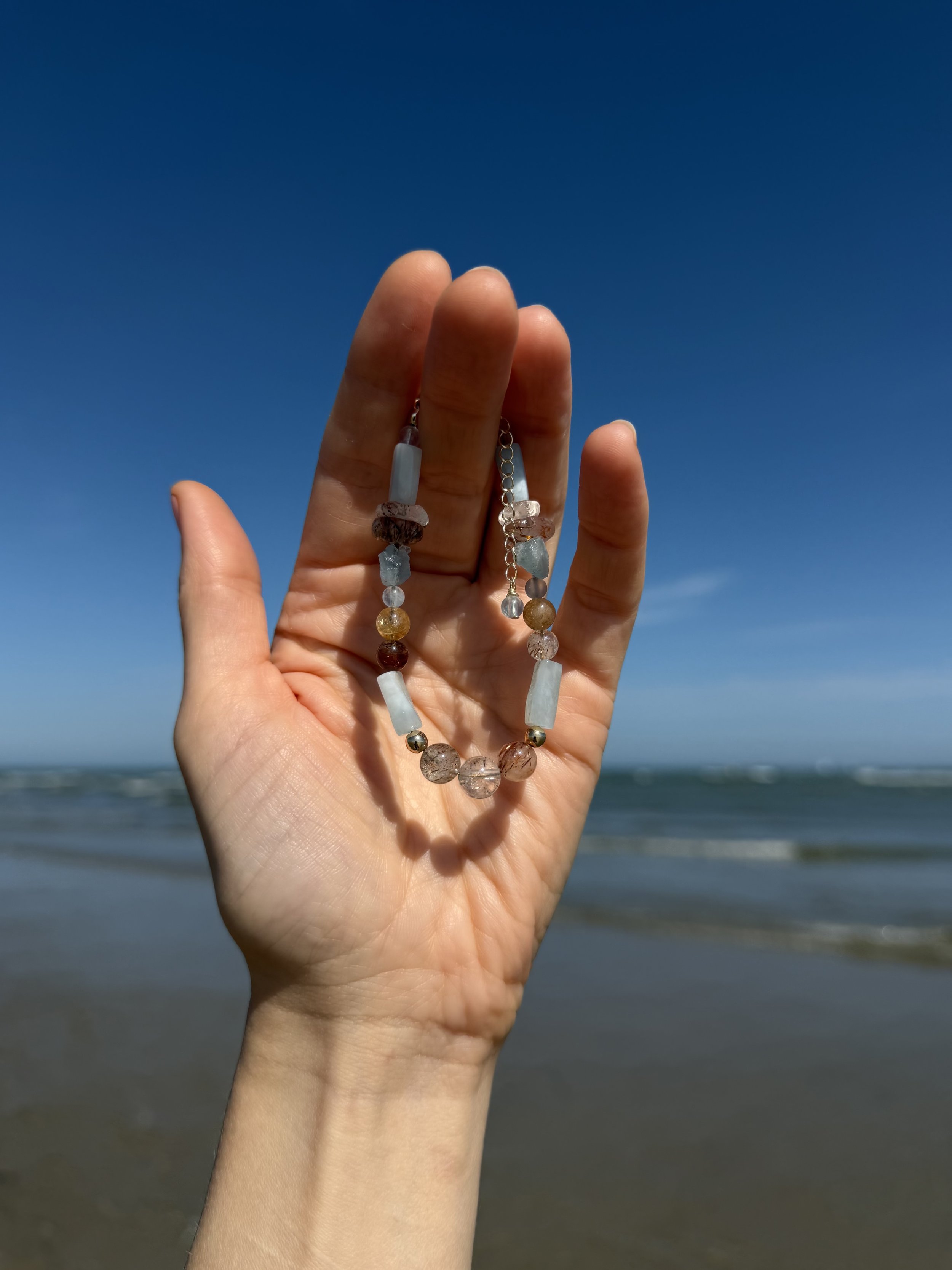 Handmade gemstone bracelet held in a hand against a blue sea and sky.