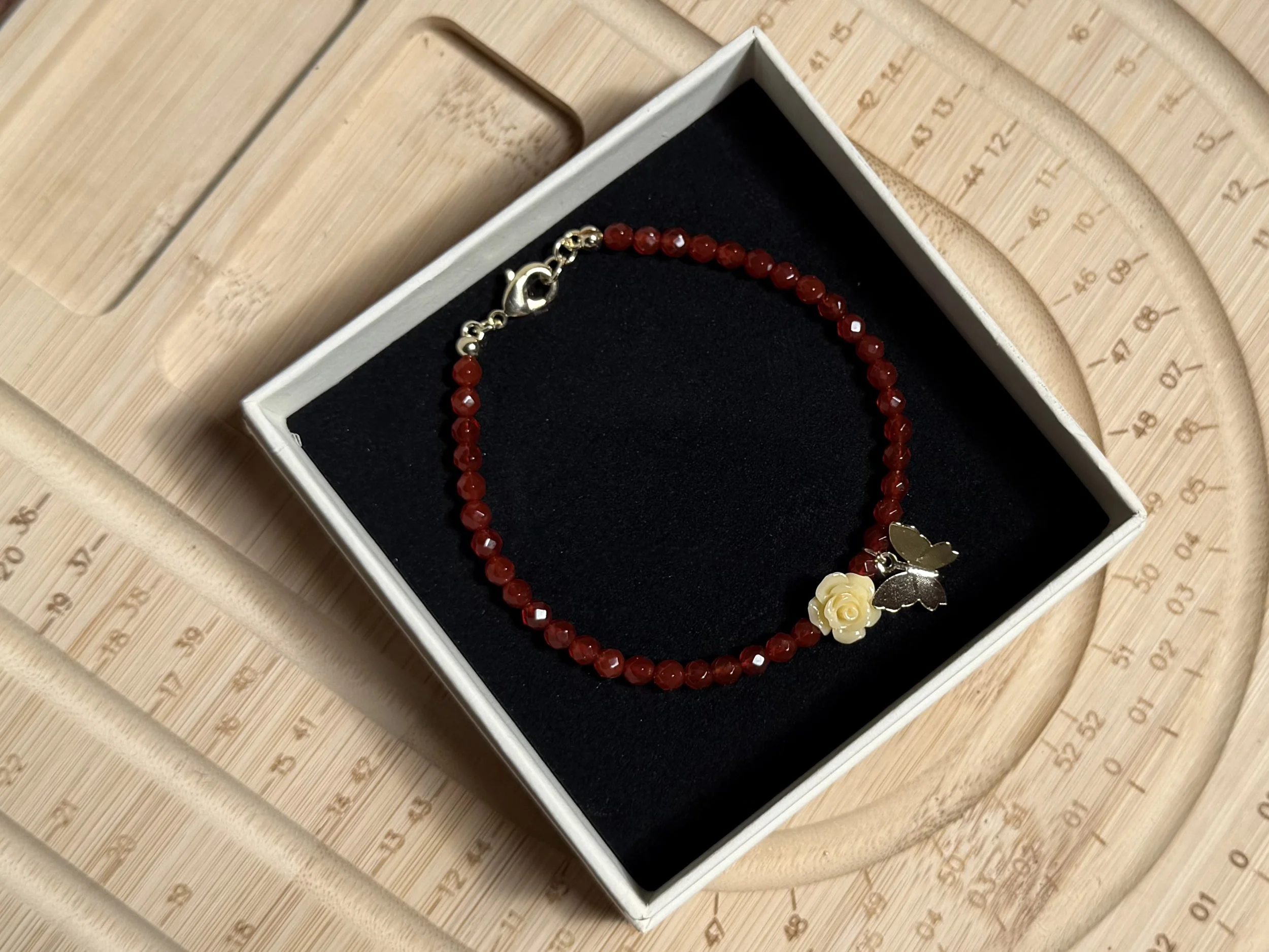 A red beaded bracelet with a gold-colored heart charm, a gold-colored flower charm, and a gold-colored butterfly charm inside a white jewelry box with a black interior, placed on a wooden surface with a circular measuring tool.
