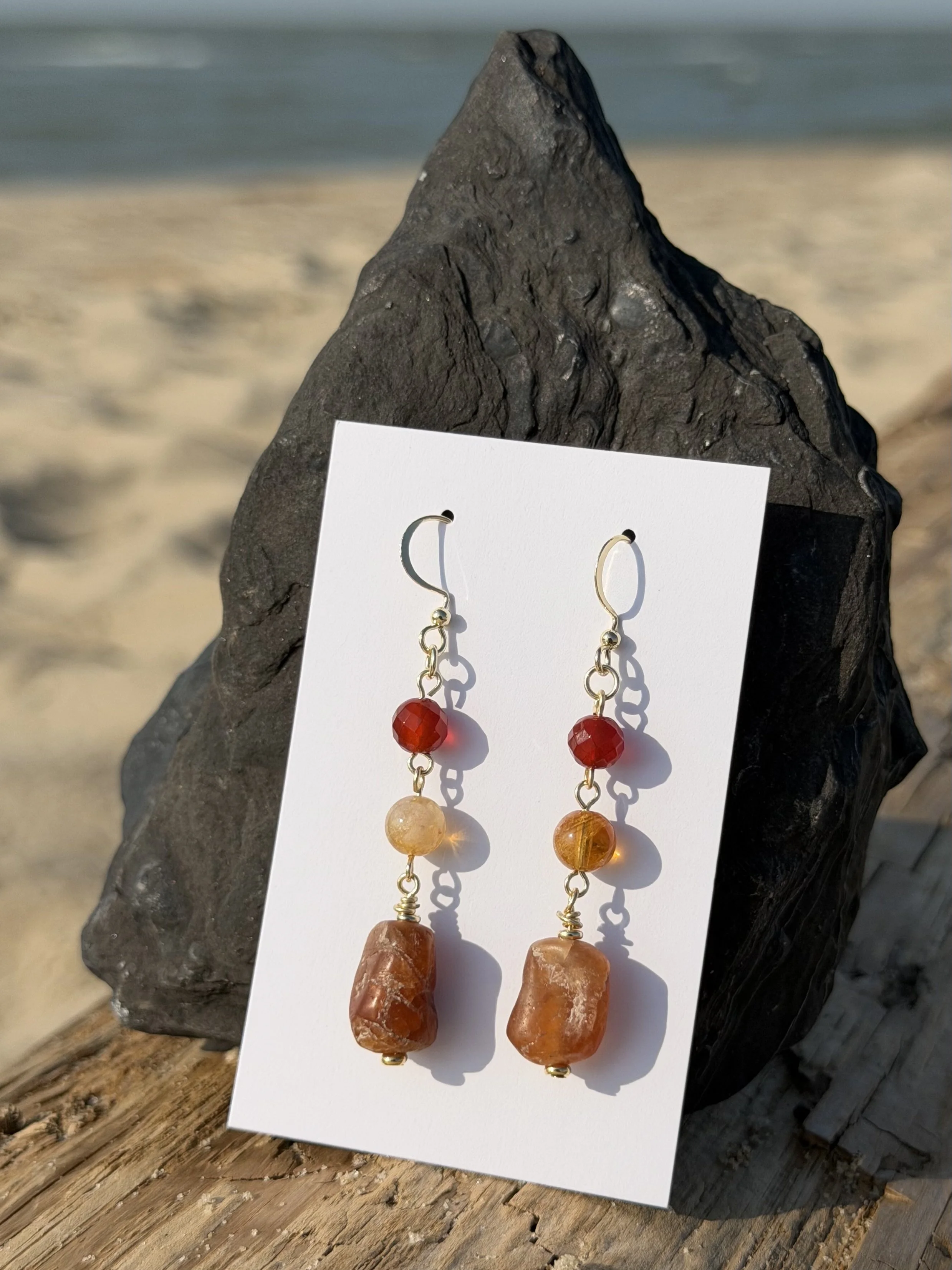 Handmade gemstone drop earrings with red, golden yellow and warm brown natural stones displayed at the beach.