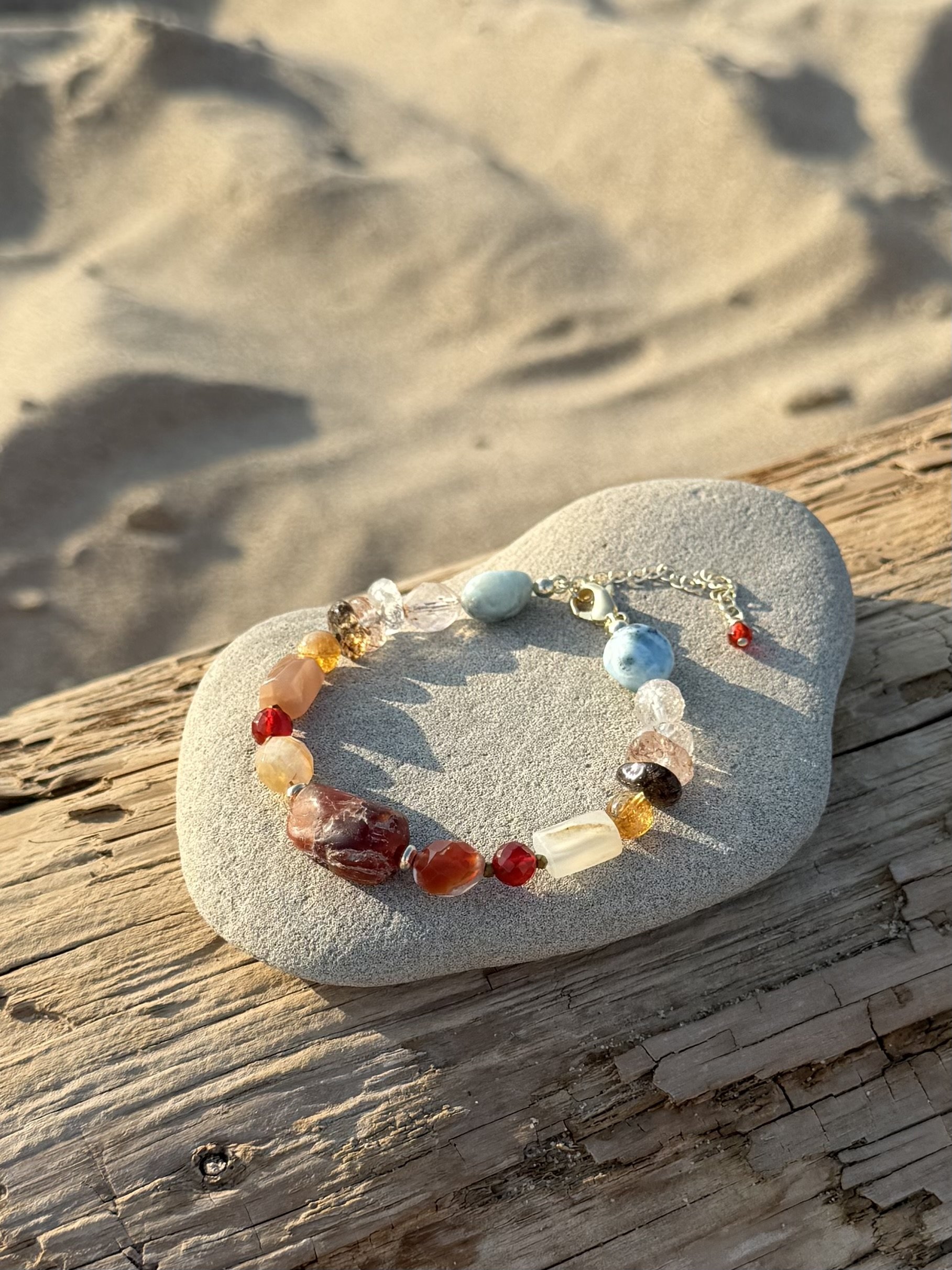 Handmade multicolour gemstone bracelet displayed on a smooth stone and driftwood in warm beach sunlight.