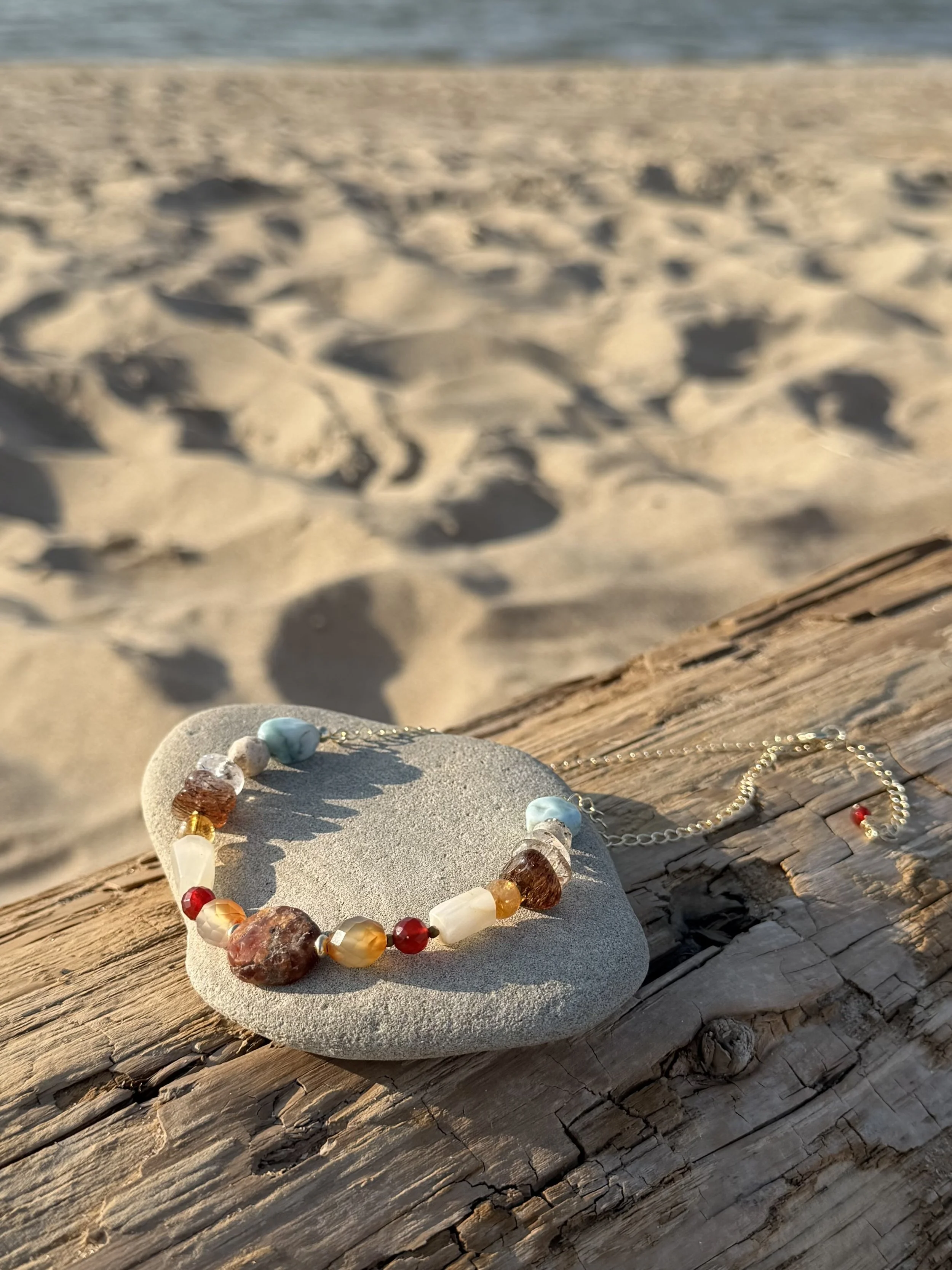 Handmade multicolour gemstone necklace displayed on a smooth stone and driftwood at the beach. stones and warm earthy tones.