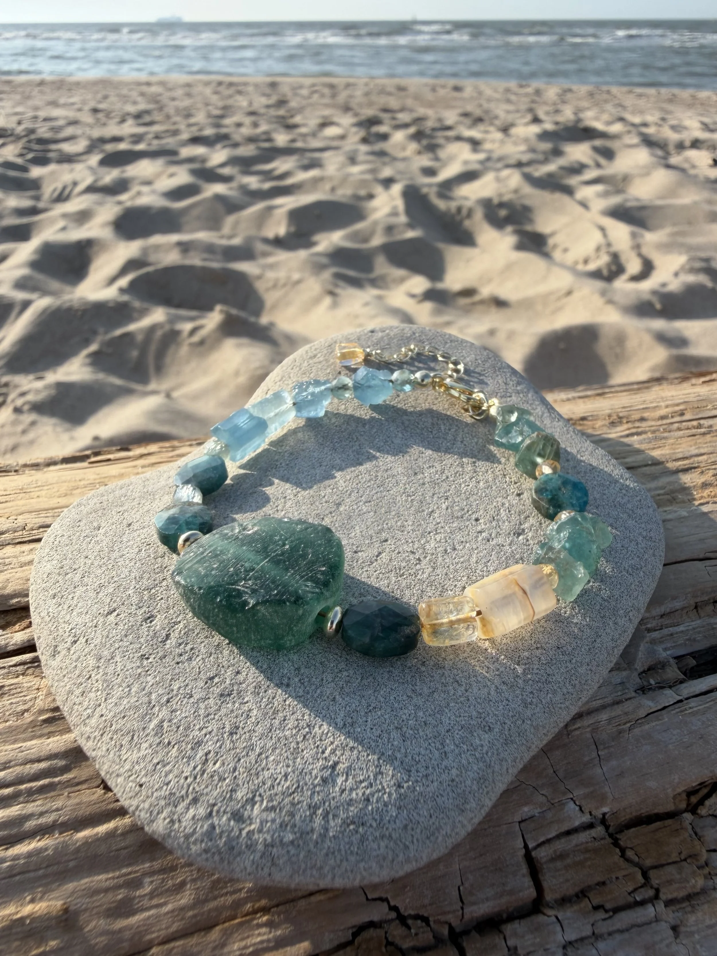 Green and blue handmade gemstone bracelet with a large natural stone centerpiece, photographed at the beach.