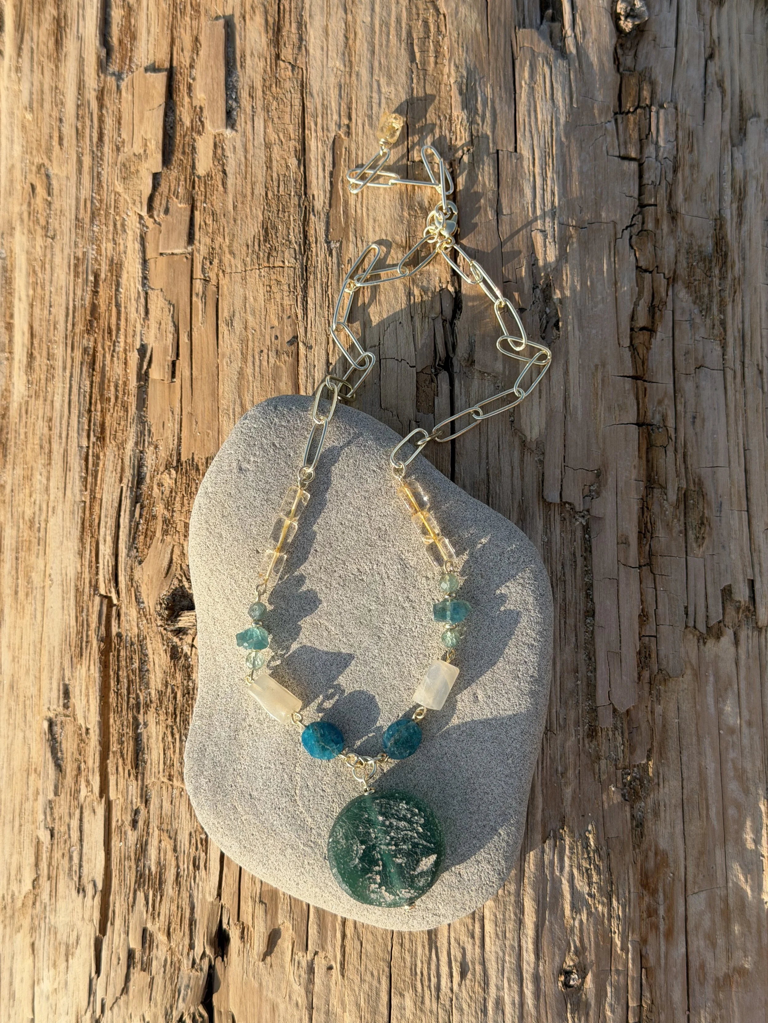 Handmade gemstone pendant necklace with green and turquoise natural stones displayed on driftwood by the sea.