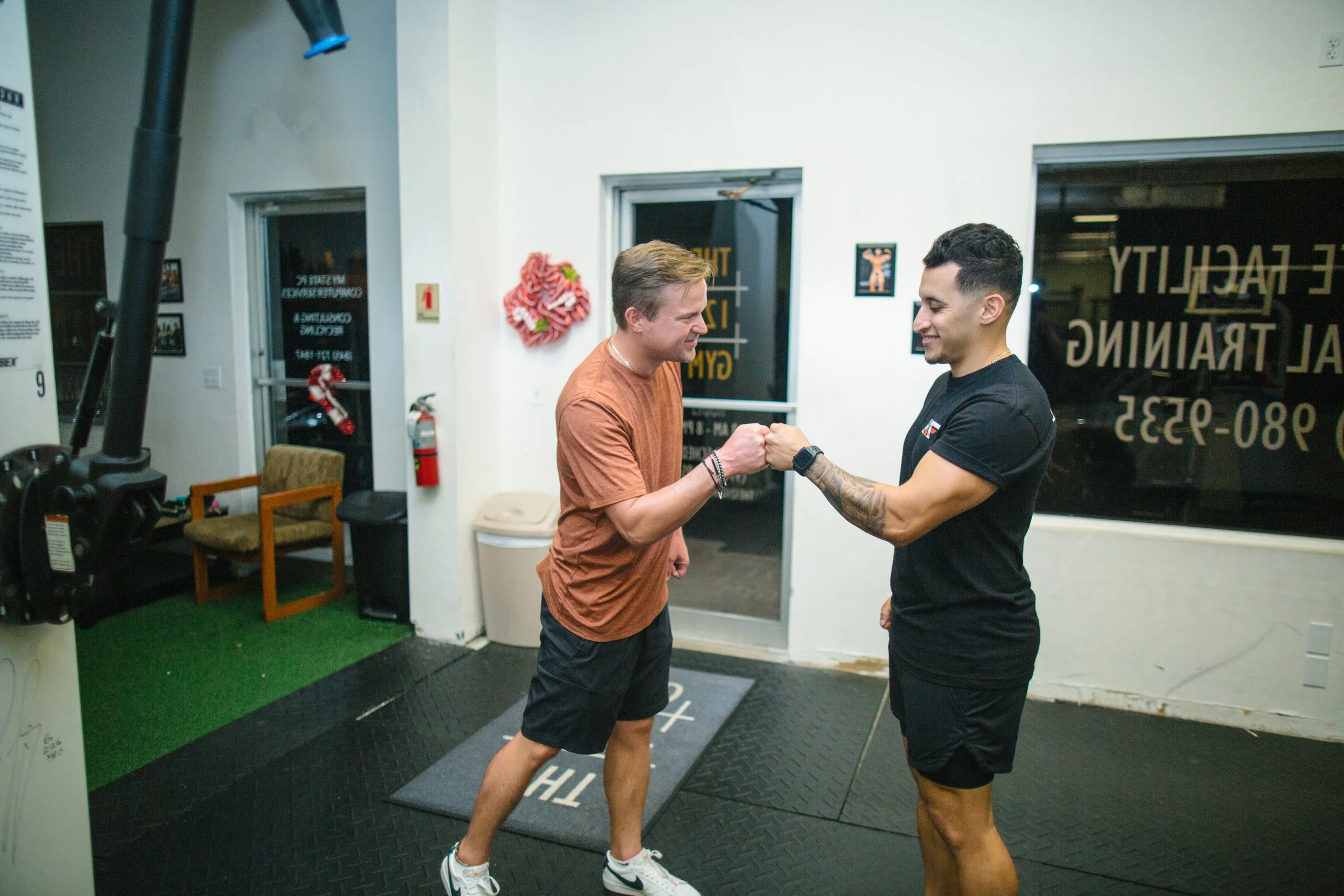 Two men are greeting each other with a fist bump in a gym or fitness studio. One is wearing a brown shirt and shorts, and the other is in black athletic wear. The background includes a door, a fire extinguisher, a framed picture, and fitness equipment.