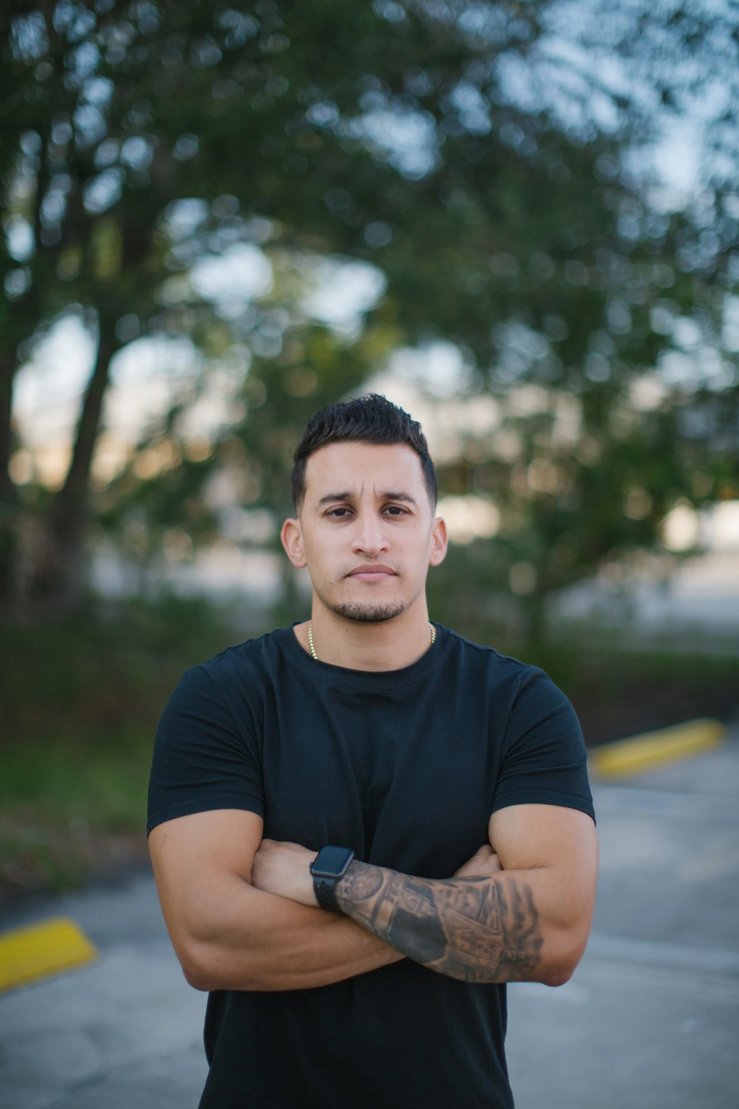 A young man with short dark hair, wearing a black t-shirt, standing outdoors with his arms crossed, background of trees and a parking lot.
