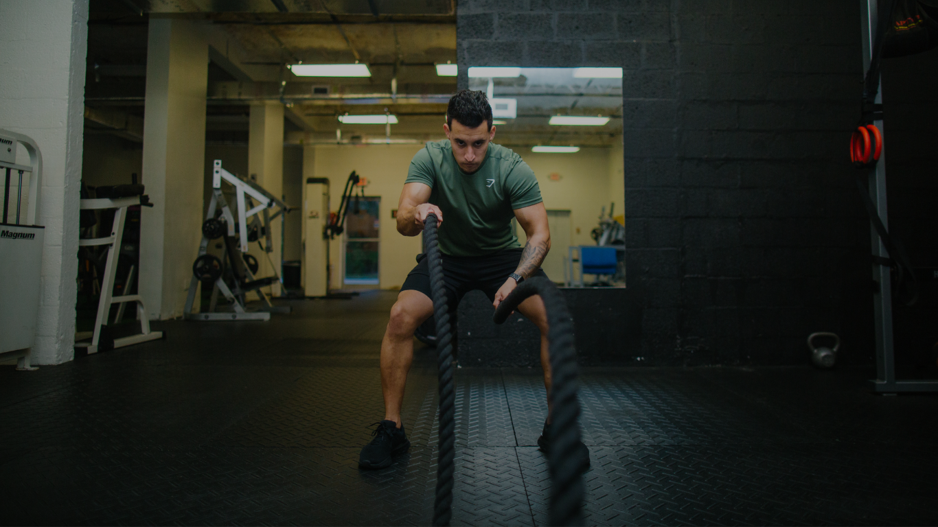 A man in a green shirt and black shorts working out with battle ropes in a gym.