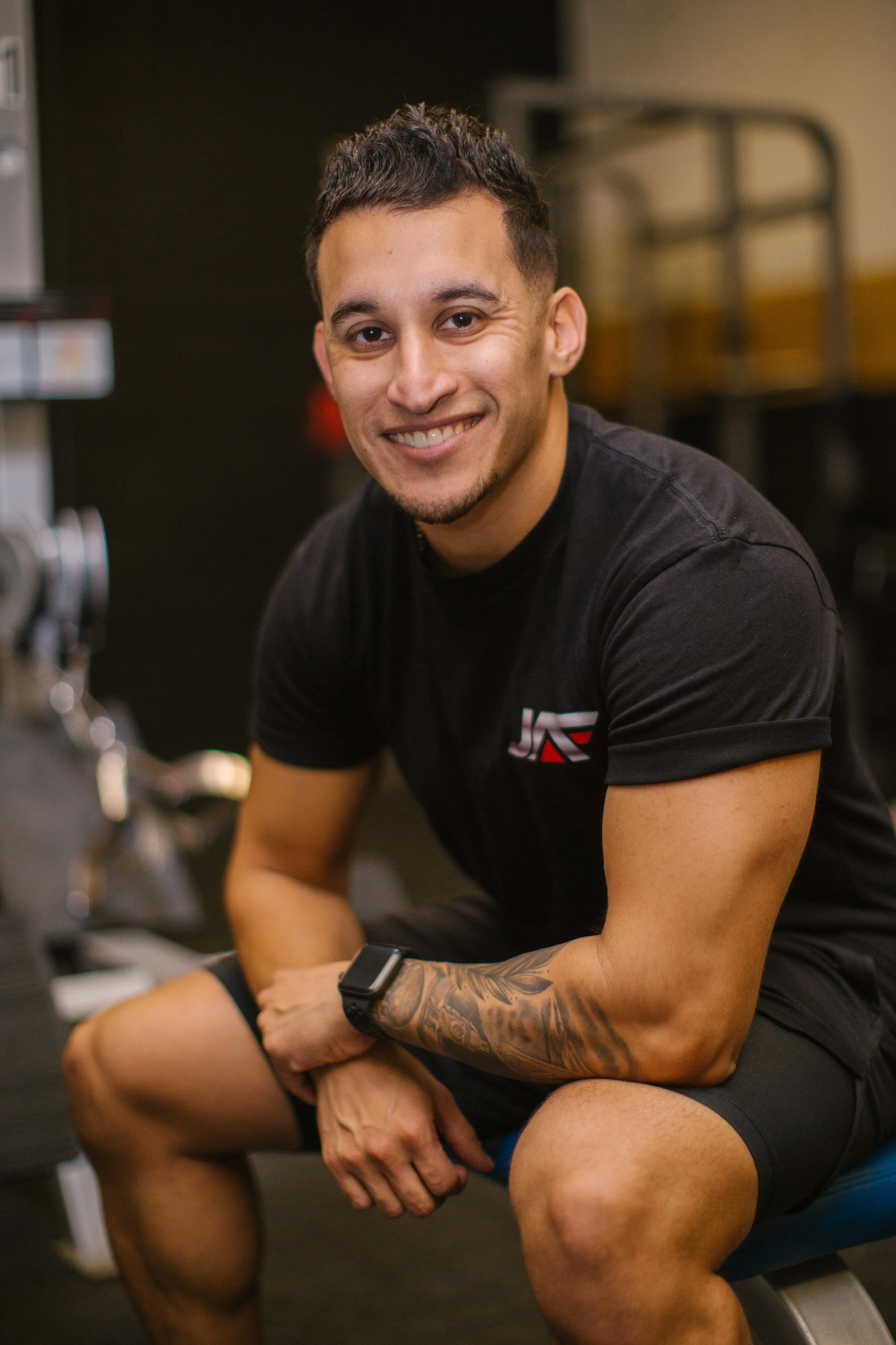 A young man with dark hair, light skin, and tattoos on his right arm, smiling at the camera, sitting on a workout bench in a gym, wearing a black T-shirt and a smartwatch.