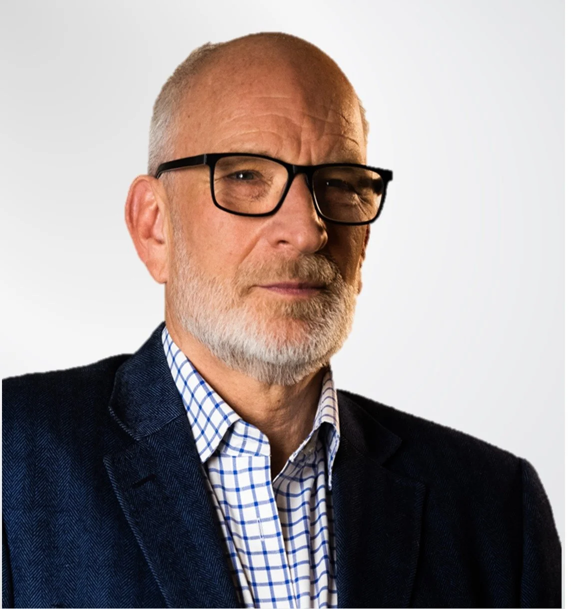Close-up portrait of an older man with glasses, a salt-and-pepper beard, dressed in a checked shirt and dark blazer against a white background.