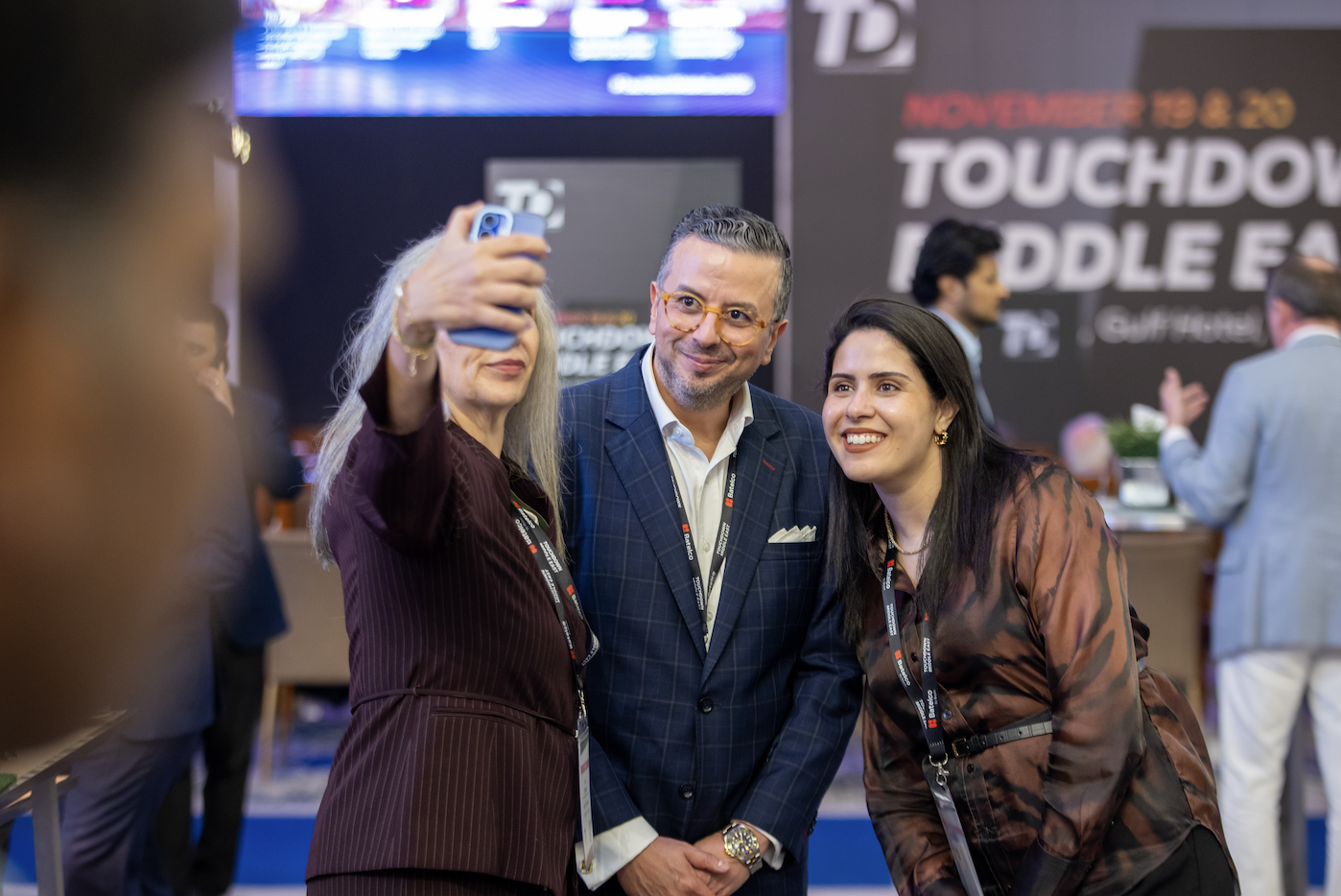 Three people taking a selfie at a conference or event, with a large screen and signage in the background reading 'November 13 & 20 Touchdown Middle East'.
