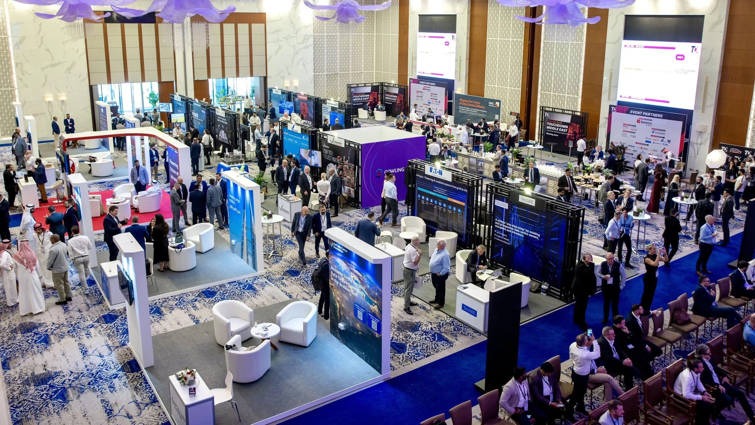 A large conference hall filled with booths, displays, and attendees. The booths feature various digital screens and banners, with people engaging in conversations and exploring exhibits.