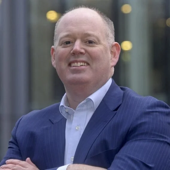 A man in a blue suit and white shirt smiling outdoors with a blurred building background and warm lighting.