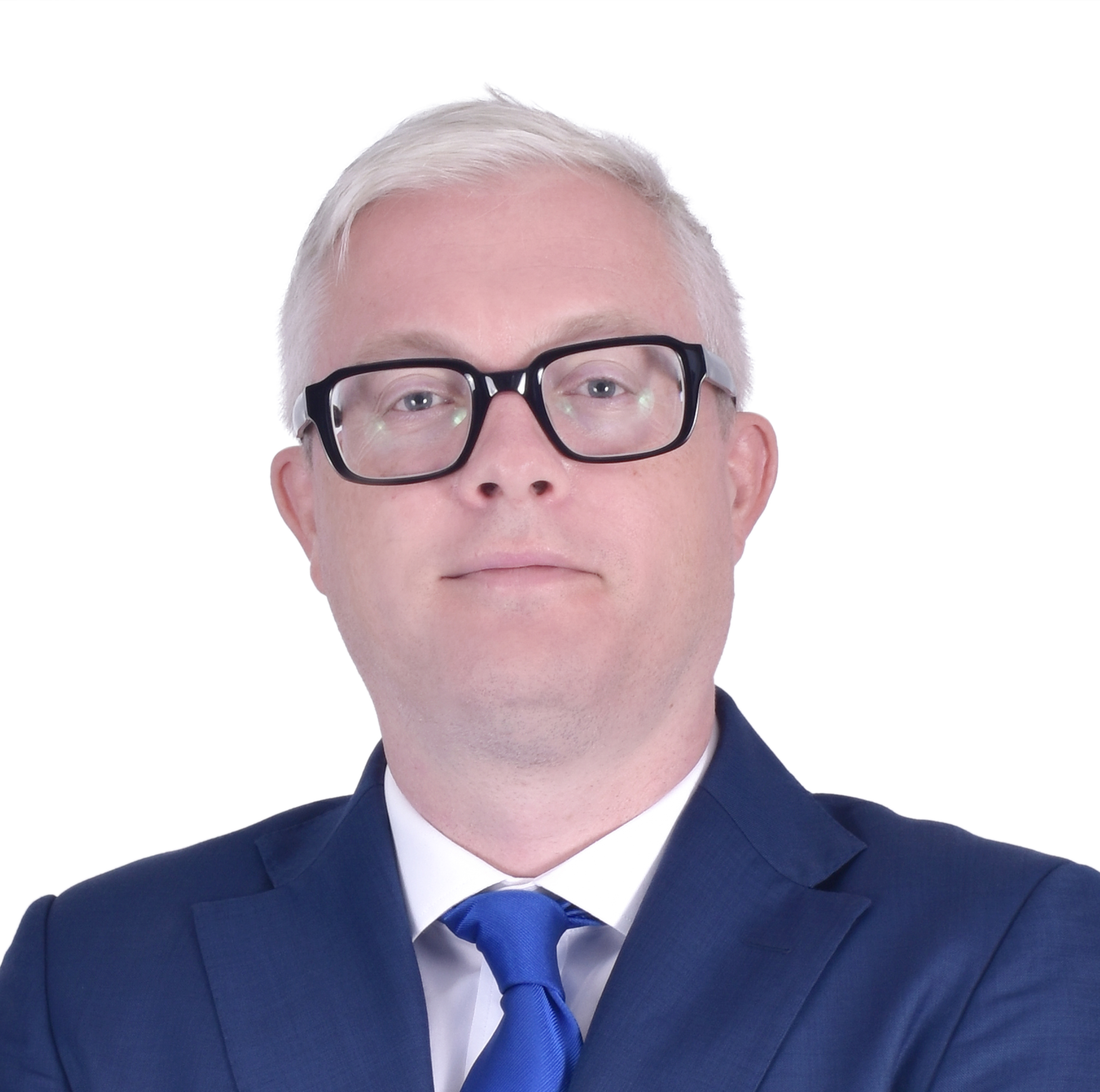Close-up of a man wearing black-rimmed glasses, a navy blue suit, white shirt, and a blue tie, with platinum blonde hair.