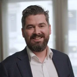 A man with dark hair and a beard, smiling, dressed in a dark blazer and white shirt, standing indoors with windows in the background.