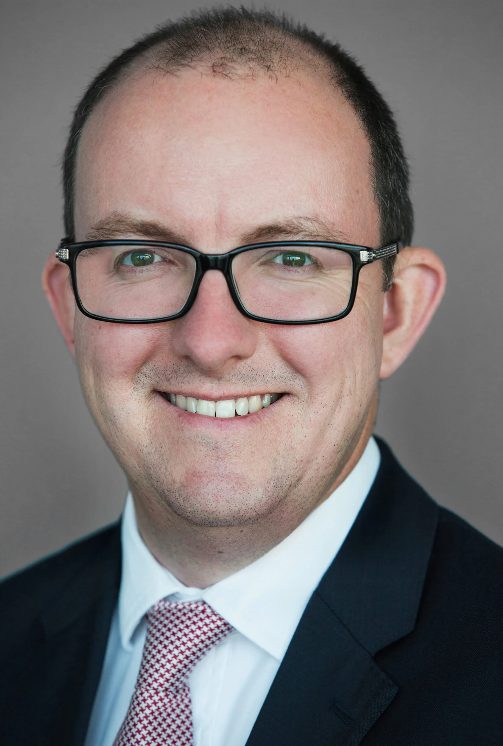 A man with short dark hair, glasses, and a wide smile, wearing a suit and red patterned tie, against a plain gray background.