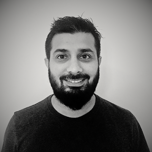 Black and white photo of a smiling man with dark hair and a beard, wearing a dark shirt, against a plain background.