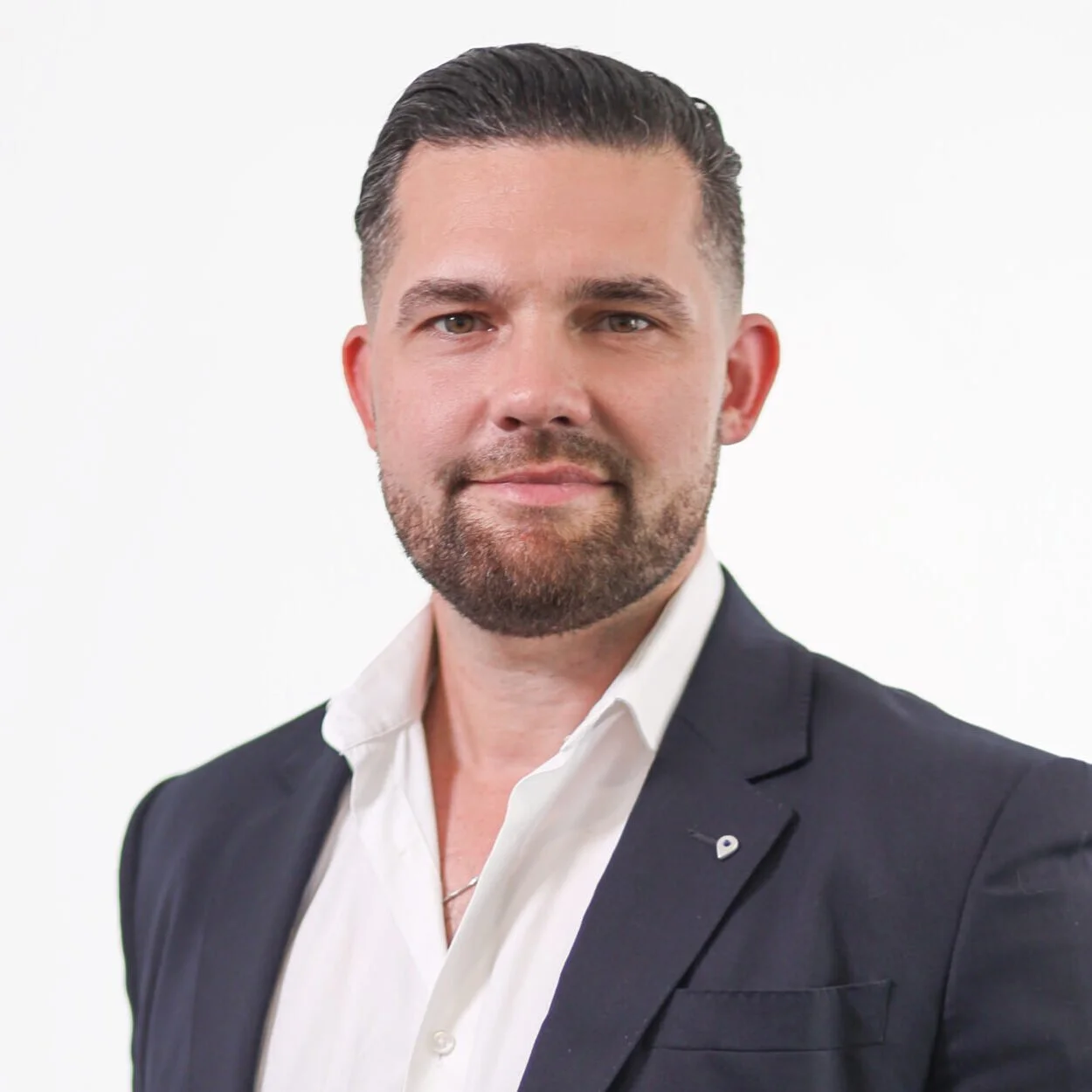 A man with short dark hair, a beard, and a mustache, wearing a dark suit jacket and a white shirt, standing against a plain white background.