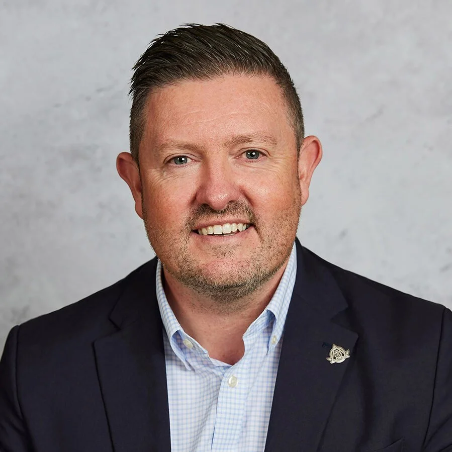 Portrait of a man with short brown hair, blue eyes, and a beard, wearing a dark blazer and a light blue checkered shirt, smiling at the camera against a gray background.